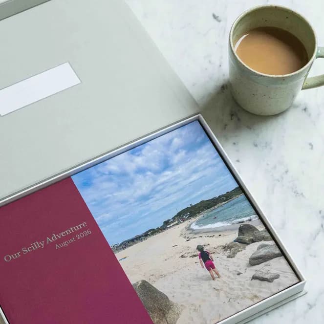A photo book labeled "Our Scilly Adventure August 2026" lies open in a box on a marble table next to a mug of tea.