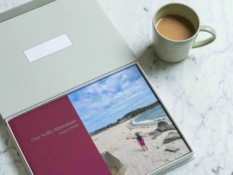 A photo book labeled "Our Scilly Adventure August 2026" lies open in a box on a marble table next to a mug of tea.