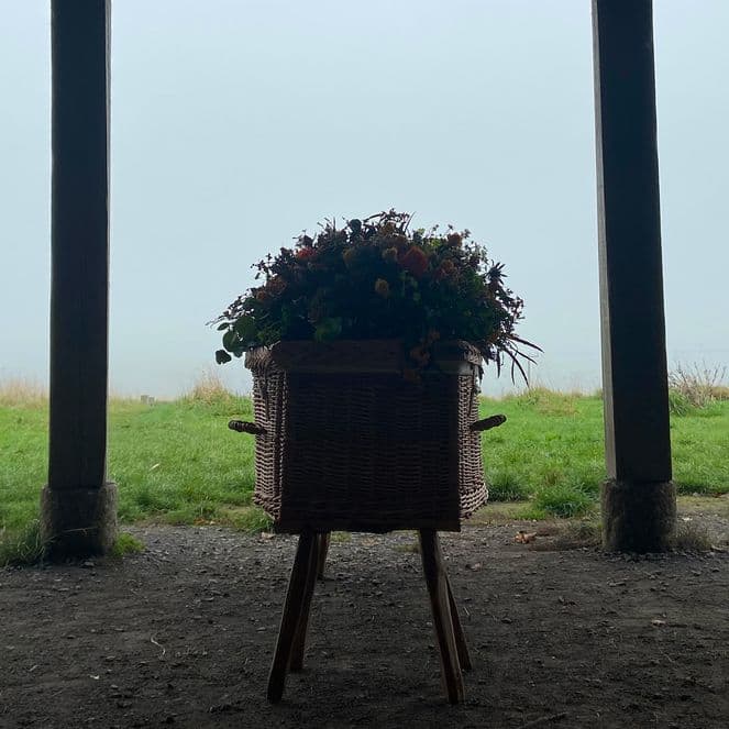 Wicker planter with flowers on a wooden stand centered between two support posts, overlooking a foggy green field.