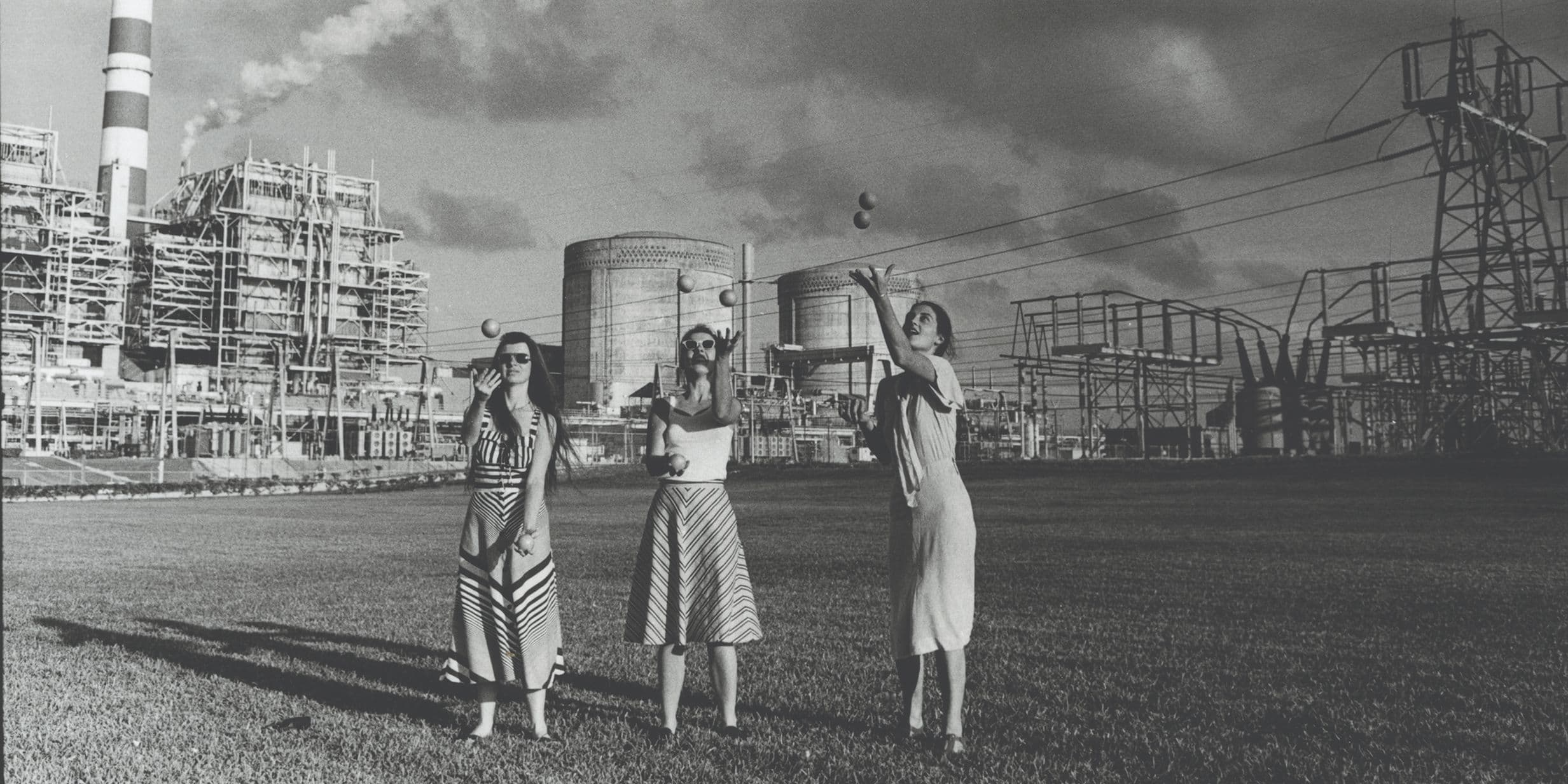Three women in dresses stand on grass before a large industrial power plant with smokestacks and electrical towers.