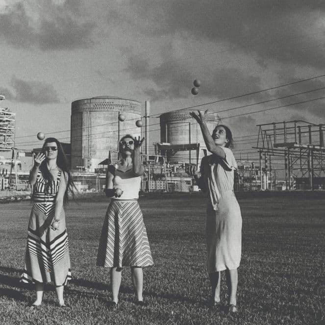 Three women in dresses stand on grass before a large industrial power plant with smokestacks and electrical towers.