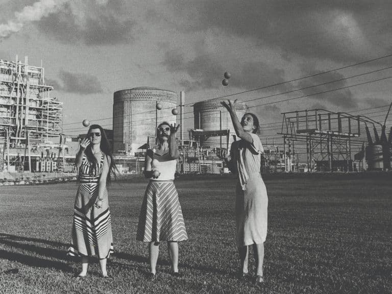 Three women in dresses stand on grass before a large industrial power plant with smokestacks and electrical towers.