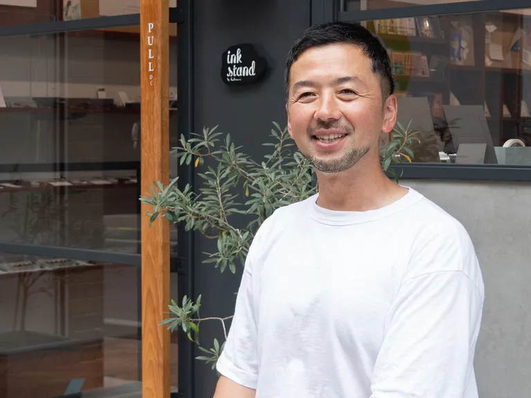 A smiling man in a white t-shirt stands outside a shop named "ink stand," with plants flanking the entrance and glass windows reflecting the interior.