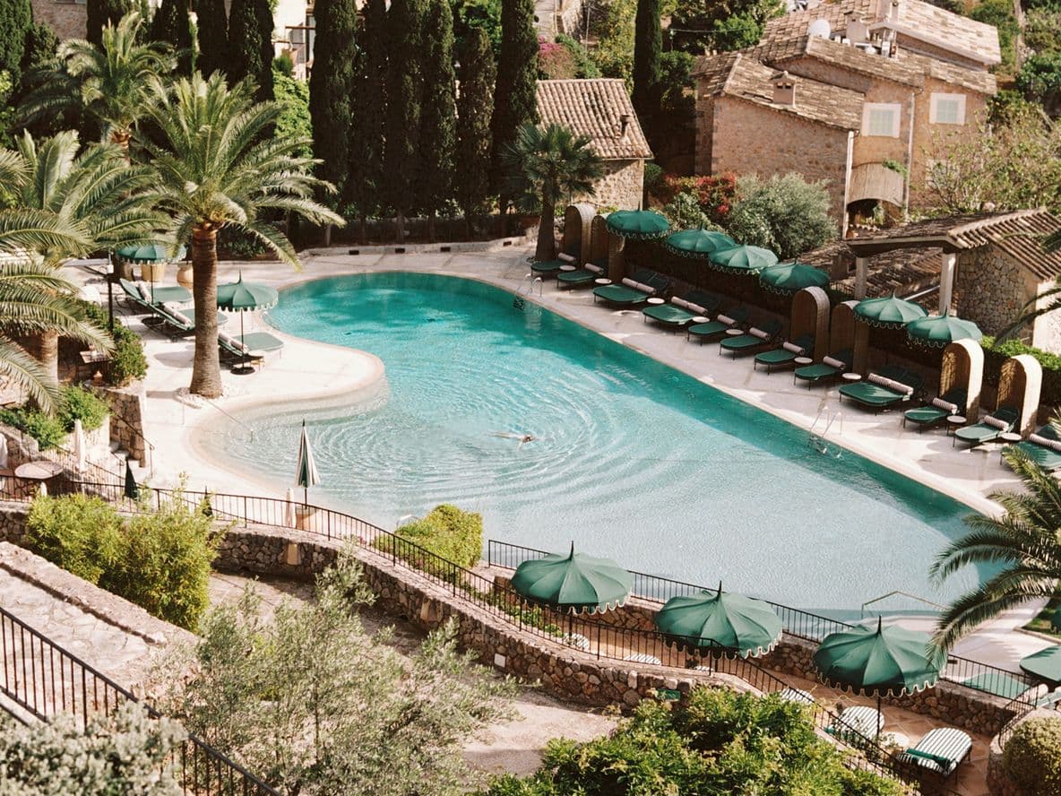 Aerial view of a serene pool surrounded by palm trees and sun loungers, with rustic stone buildings in the background.