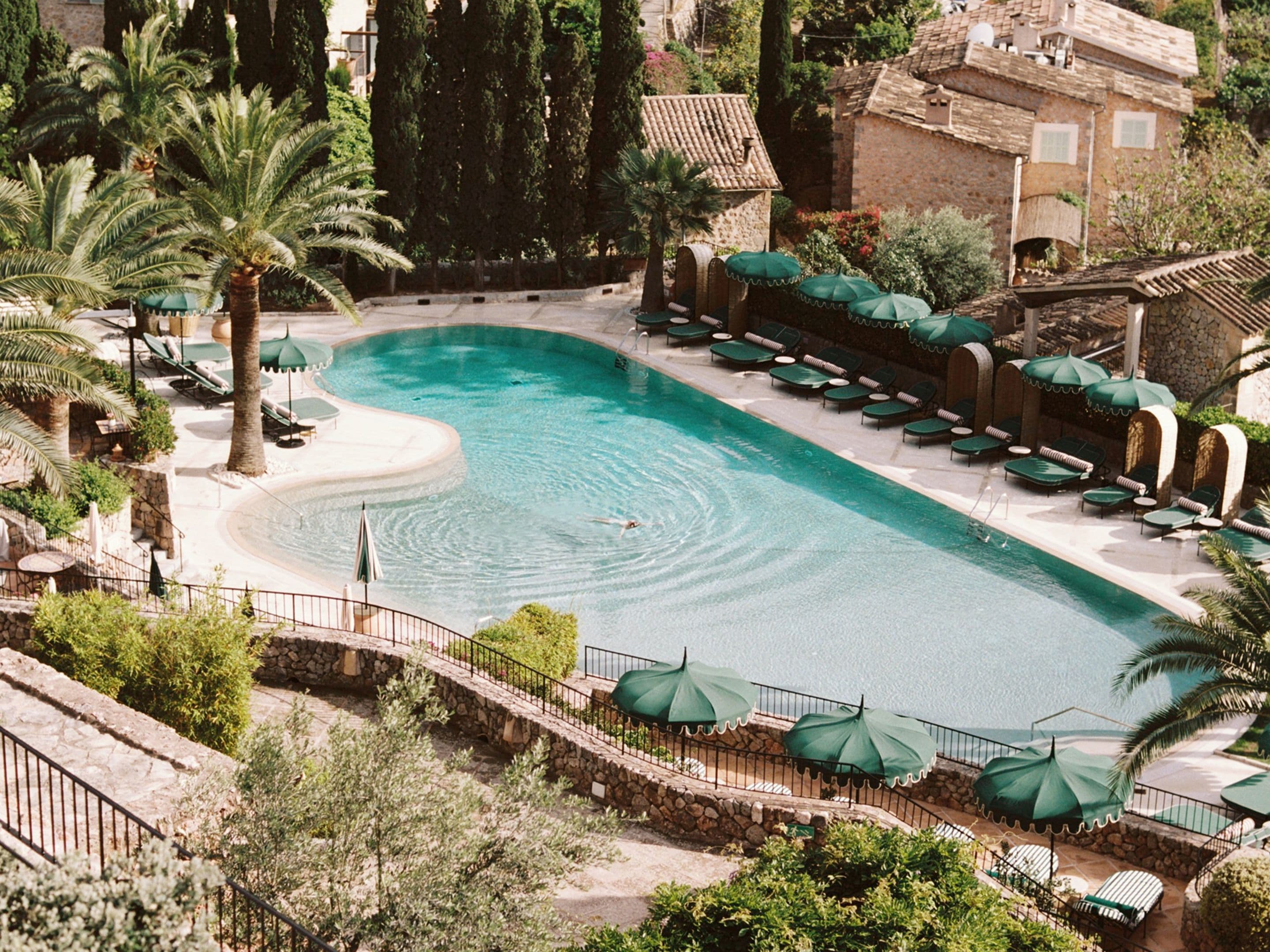 Aerial view of a serene pool surrounded by palm trees and sun loungers, with rustic stone buildings in the background.