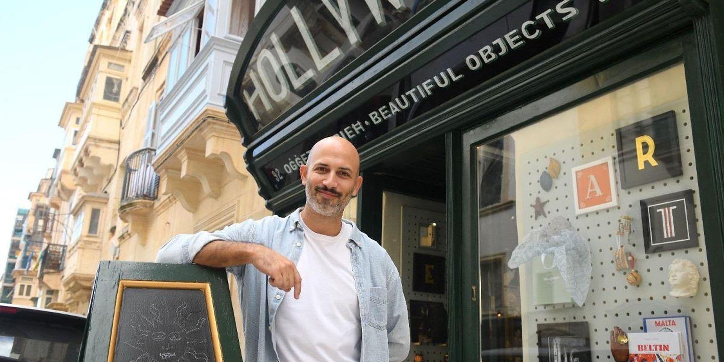 A man stands smiling outside a store named "Hollywood" with a chalkboard sign and display window showing various decorative items.