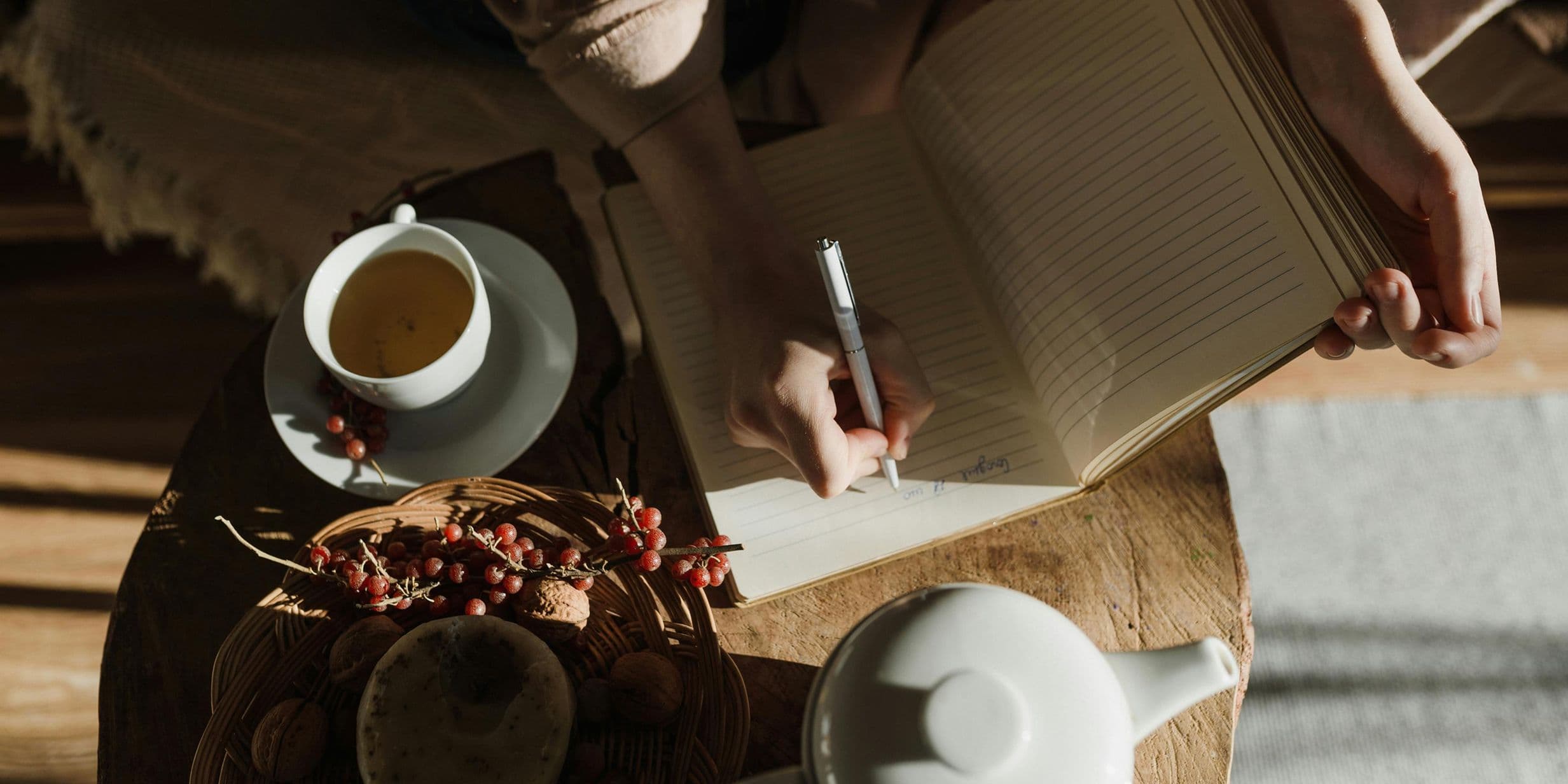 Person writing in a notebook at a table with a cup of tea, a teapot, and a basket of berries in warm, natural light.