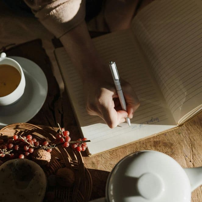 Person writing in a notebook at a table with a cup of tea, a teapot, and a basket of berries in warm, natural light.