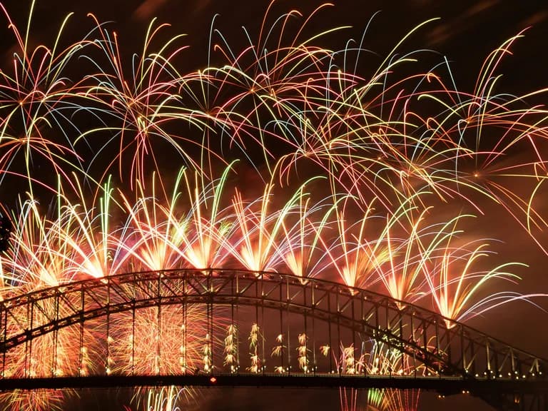 Colorful fireworks light up the night sky over a large, arched bridge silhouetted against glowing bursts.
