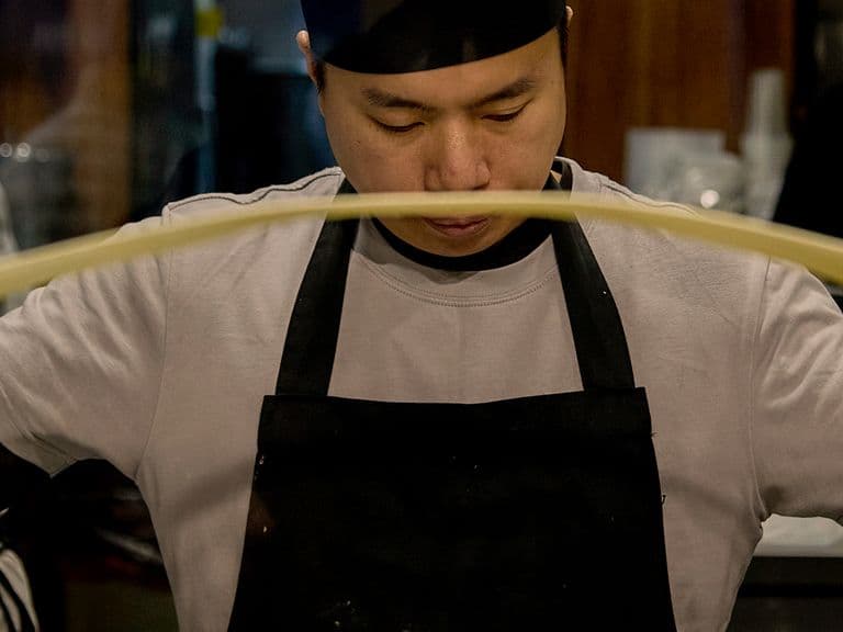 A chef in a black apron and hat stretches dough.