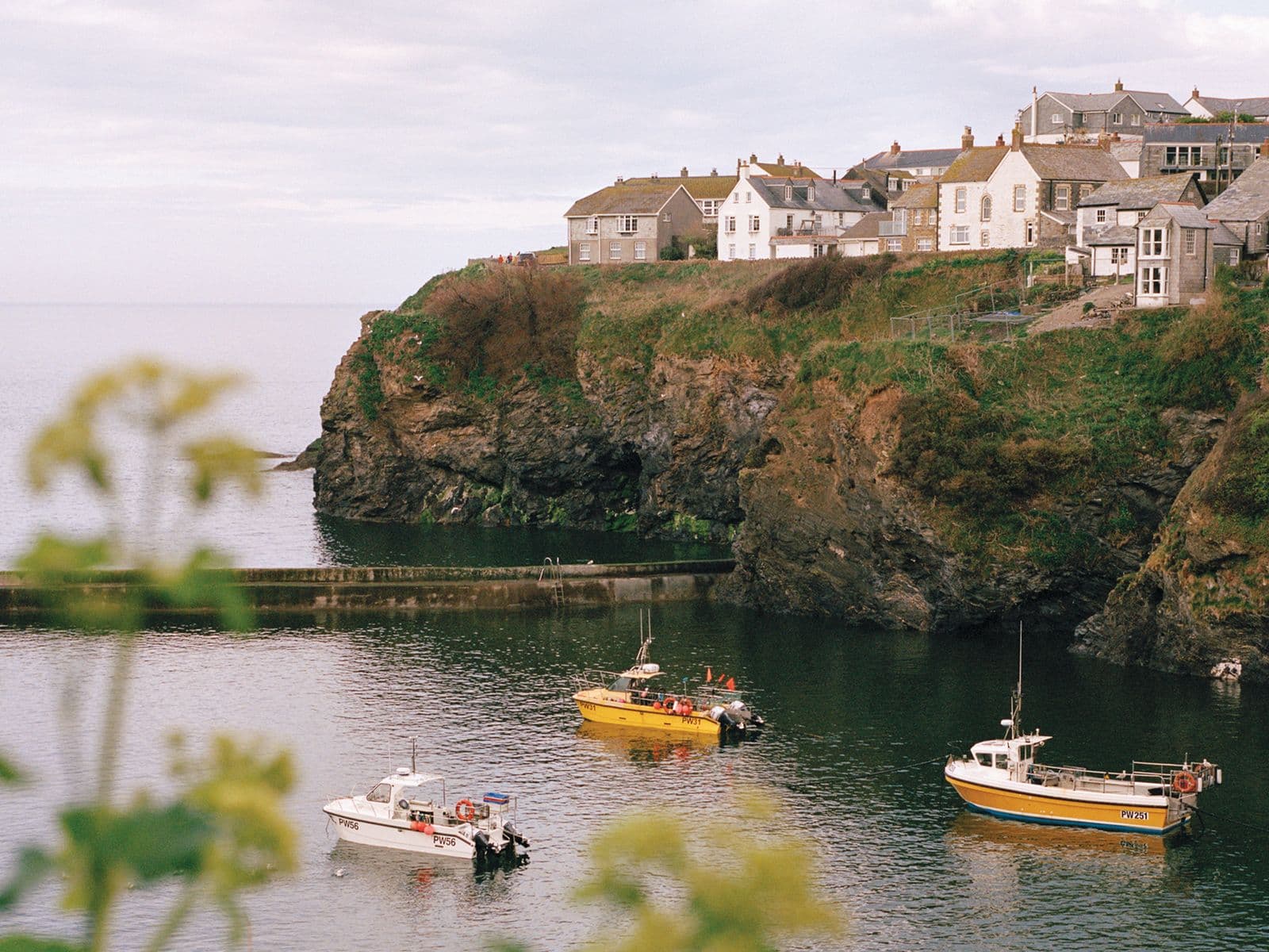 Coastal village with houses on a cliff, overlooking a calm sea with three small boats. Foreground features blurred greenery.
