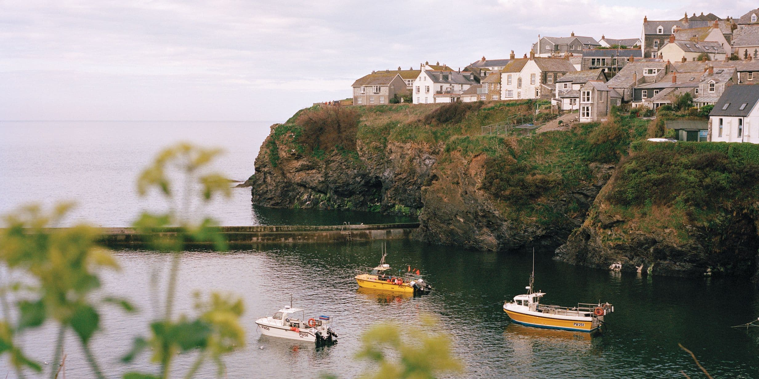 Coastal village with houses on a cliff, overlooking a calm sea with three small boats. Foreground features blurred greenery.