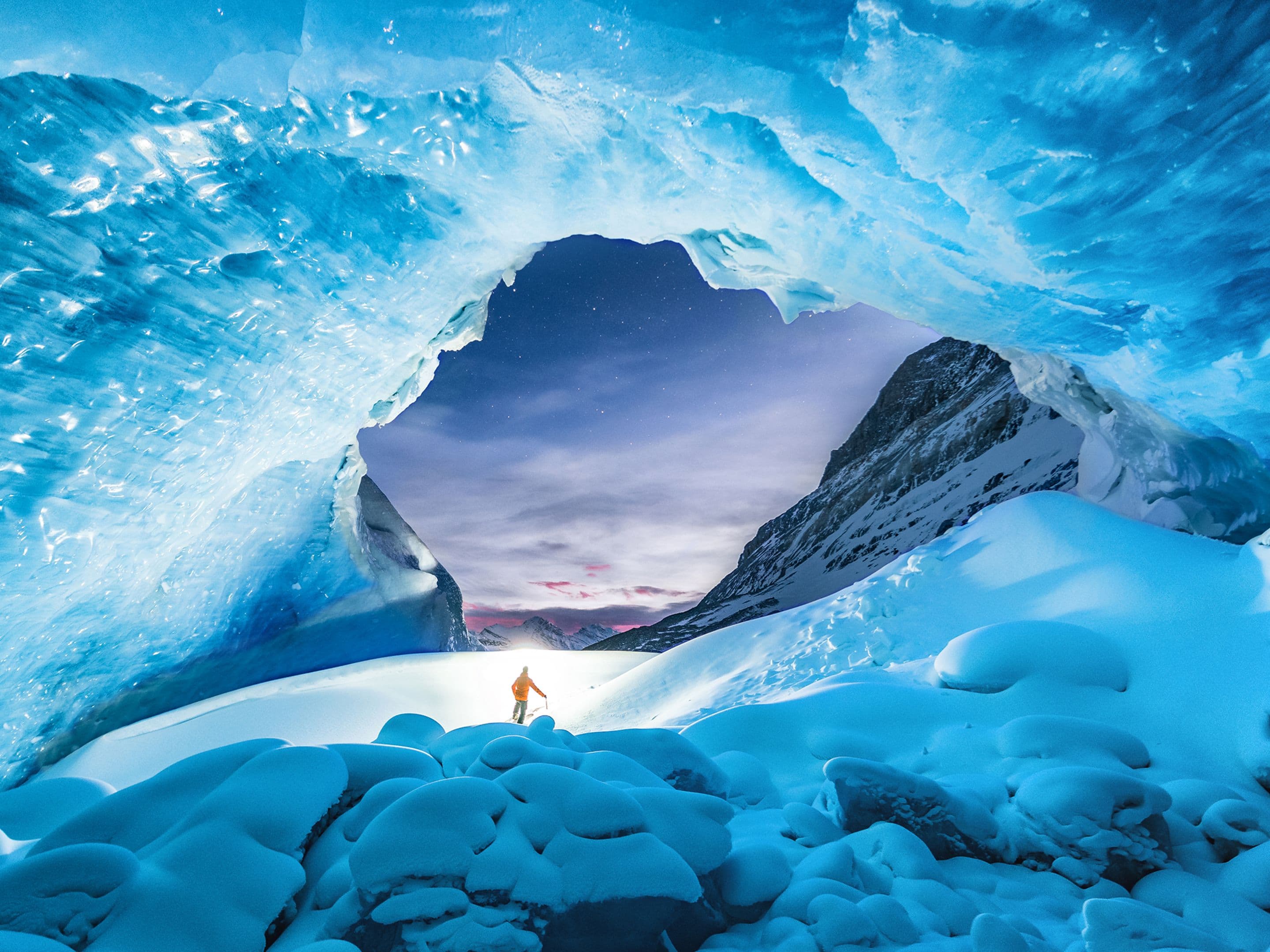 Person in winter clothing stands under a vast, blue ice cave with a view of snowy mountains and a twilight sky in the background.