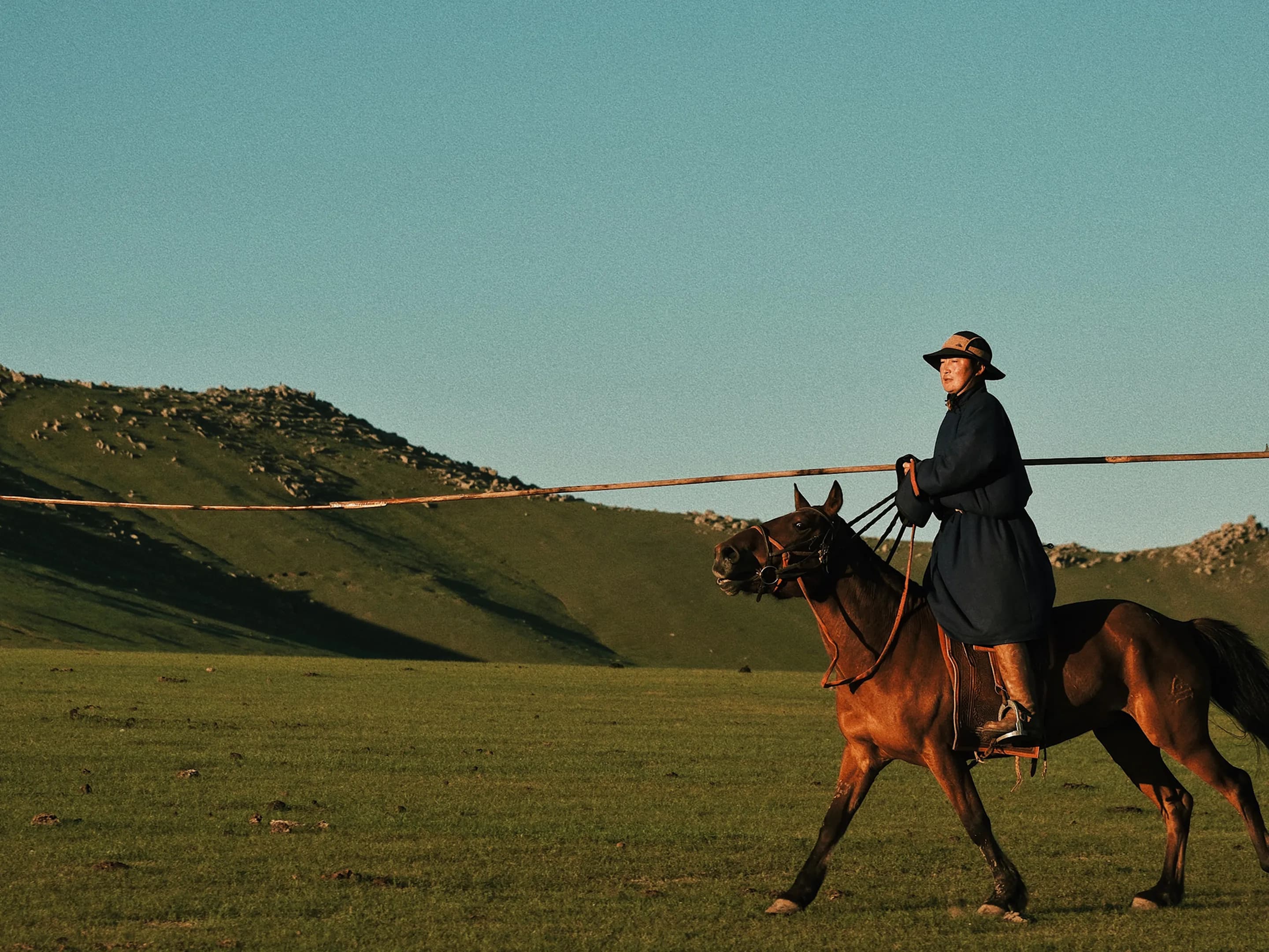 Person in a dark coat and hat rides a brown horse across a vast, grassy landscape with distant hills under a clear blue sky.