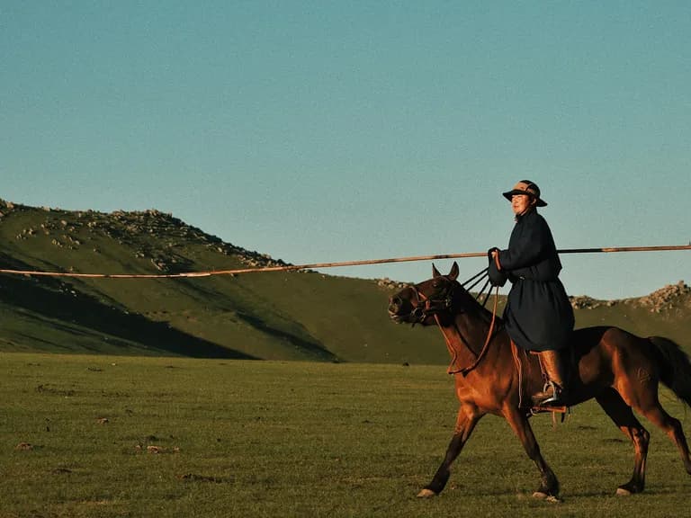 Person in a dark coat and hat rides a brown horse across a vast, grassy landscape with distant hills under a clear blue sky.