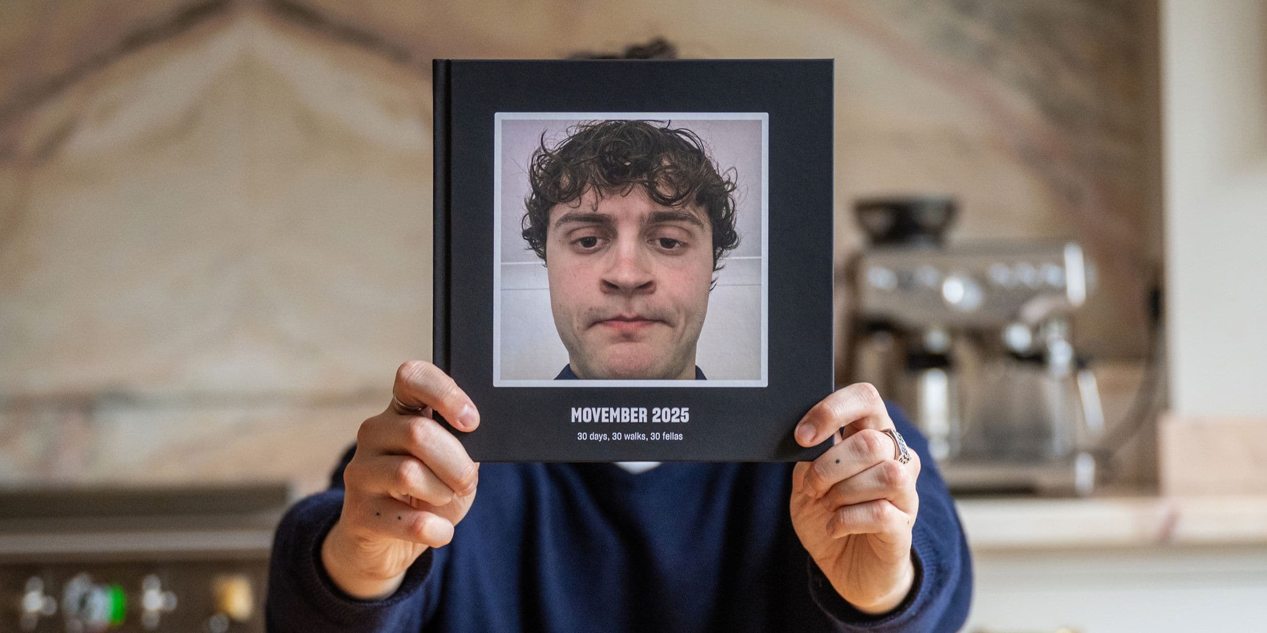 Person holding a photo album titled "Movember 2023" featuring a close-up of a man with curly hair. Kitchen setting with a coffee machine.