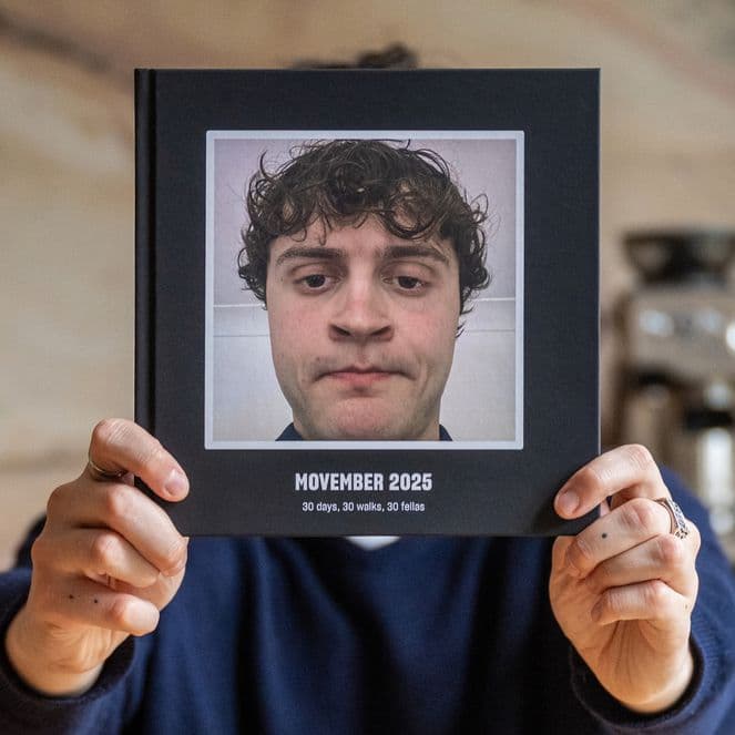 Person holding a photo album titled "Movember 2023" featuring a close-up of a man with curly hair. Kitchen setting with a coffee machine.