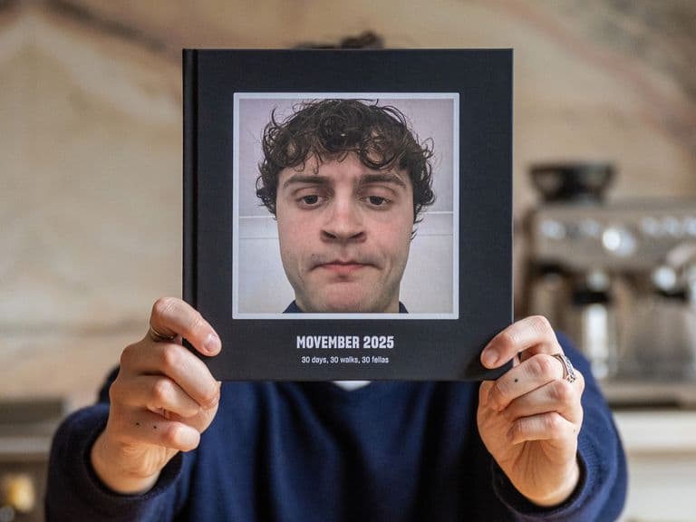 Person holding a photo album titled "Movember 2023" featuring a close-up of a man with curly hair. Kitchen setting with a coffee machine.
