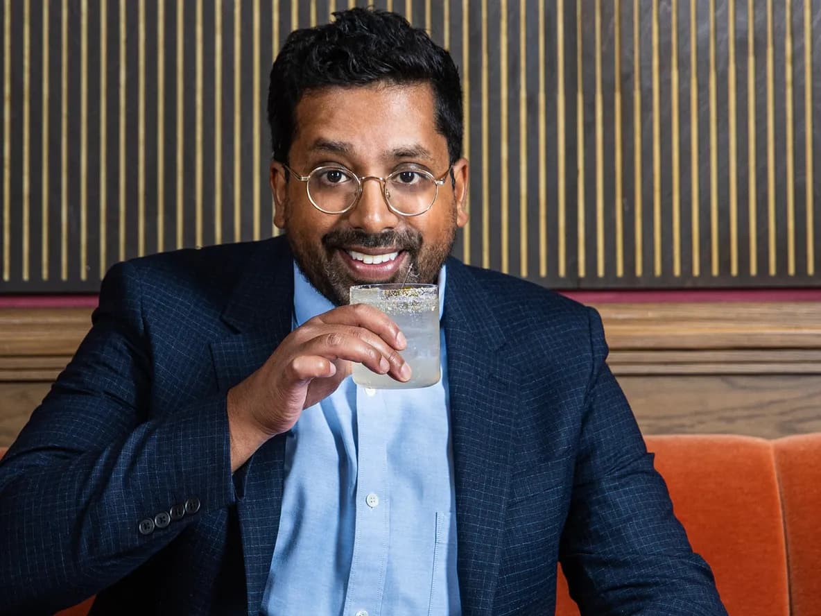 Man in a suit sitting on an orange bench, smiling, and sipping a drink from a glass with a salted rim.