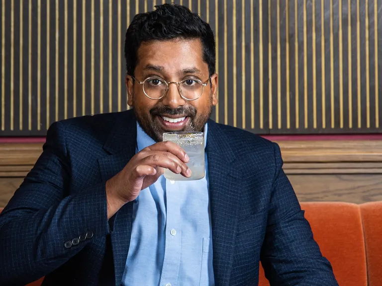 Man in a suit sitting on an orange bench, smiling, and sipping a drink from a glass with a salted rim.