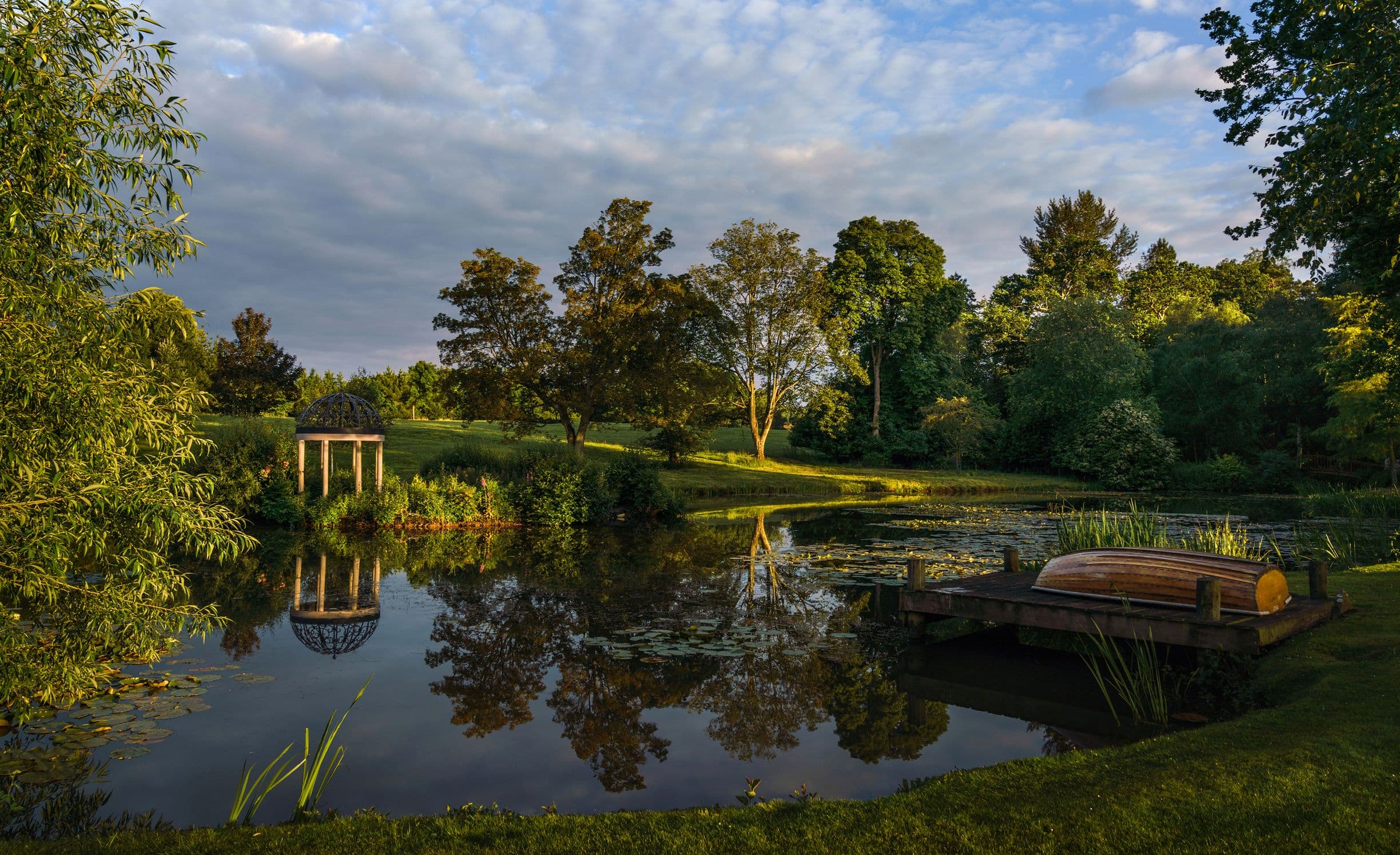 Serene pond with a wooden dock, a small boat, and a gazebo. Surrounded by lush trees under a partly cloudy sky.