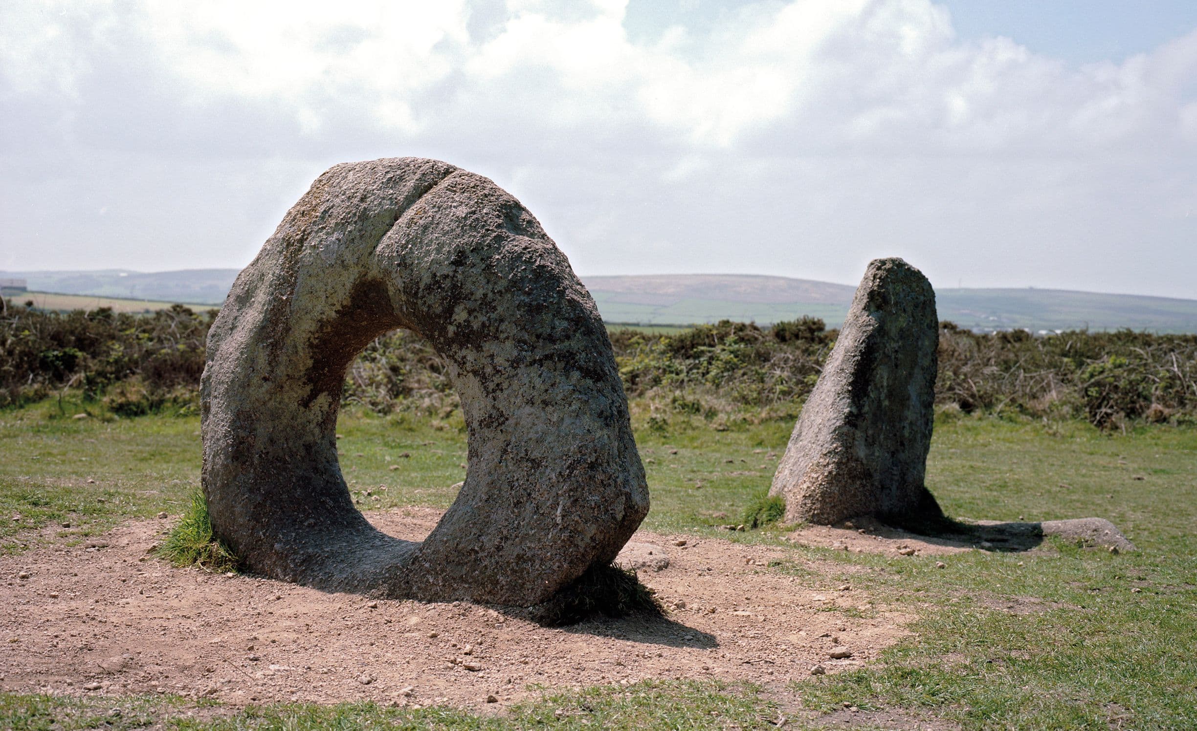 Two ancient standing stones on a grassy landscape, one with a circular hole, under a partly cloudy sky.