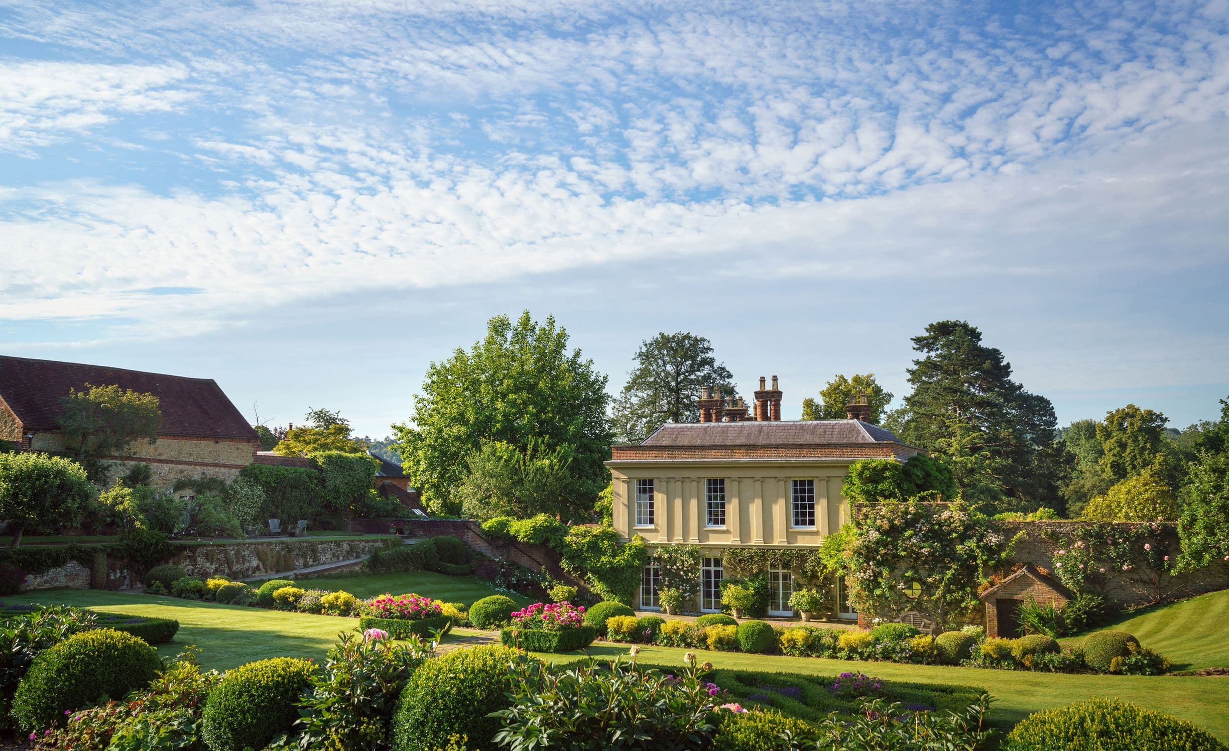 A picturesque countryside scene with a charming house surrounded by lush gardens, manicured lawns, and a clear blue sky with scattered clouds.