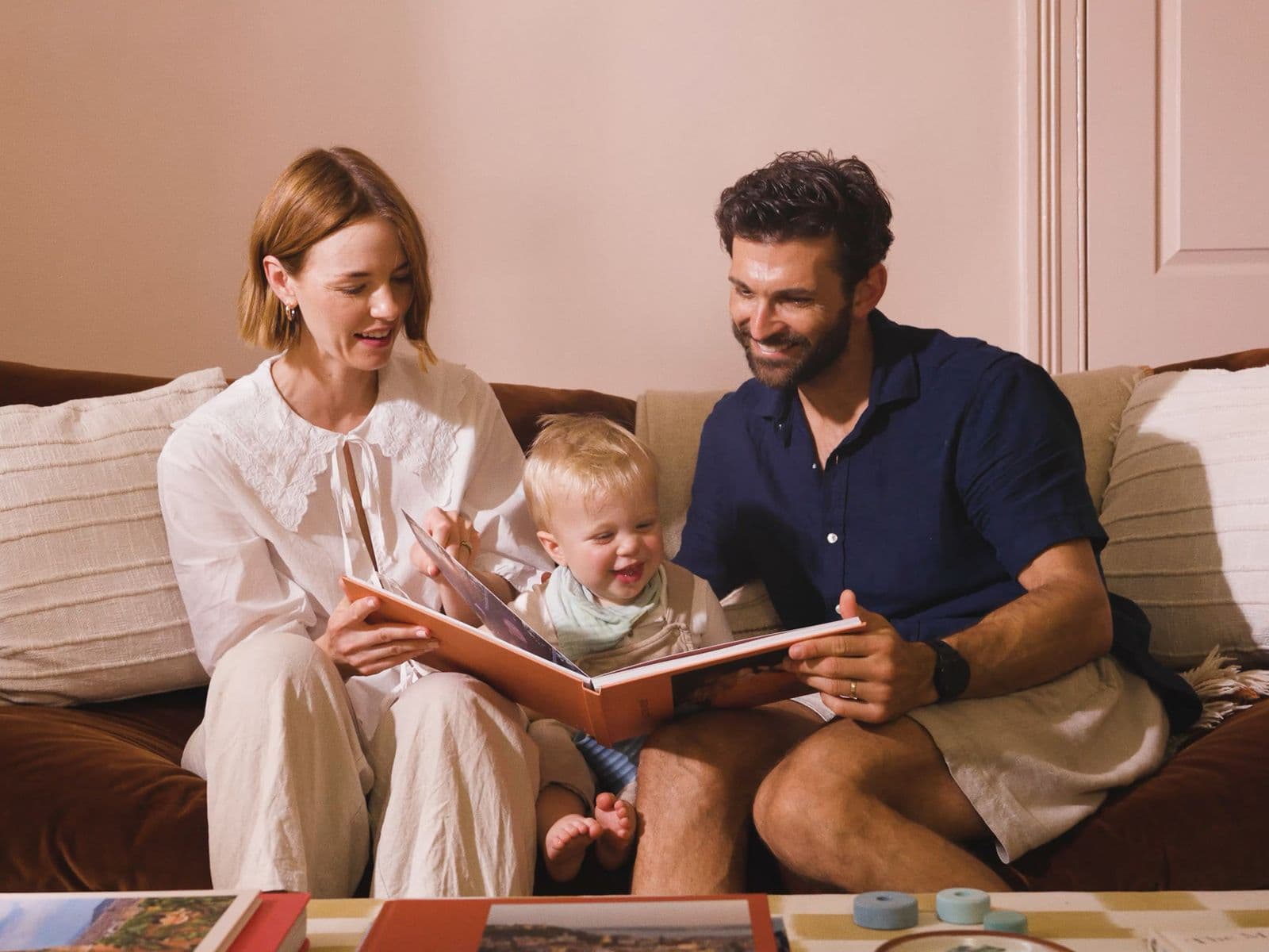 A happy couple look through a baby photo book with a toddler sat between them on a sofa