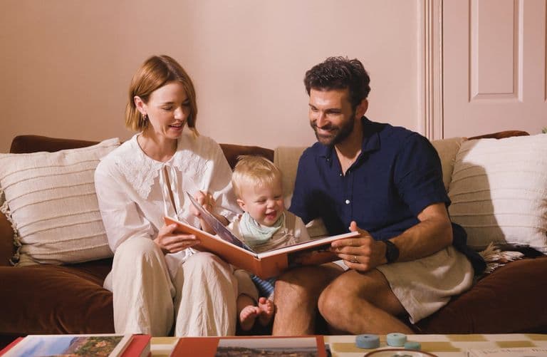 A happy couple look through a baby photo book with a toddler sat between them on a sofa