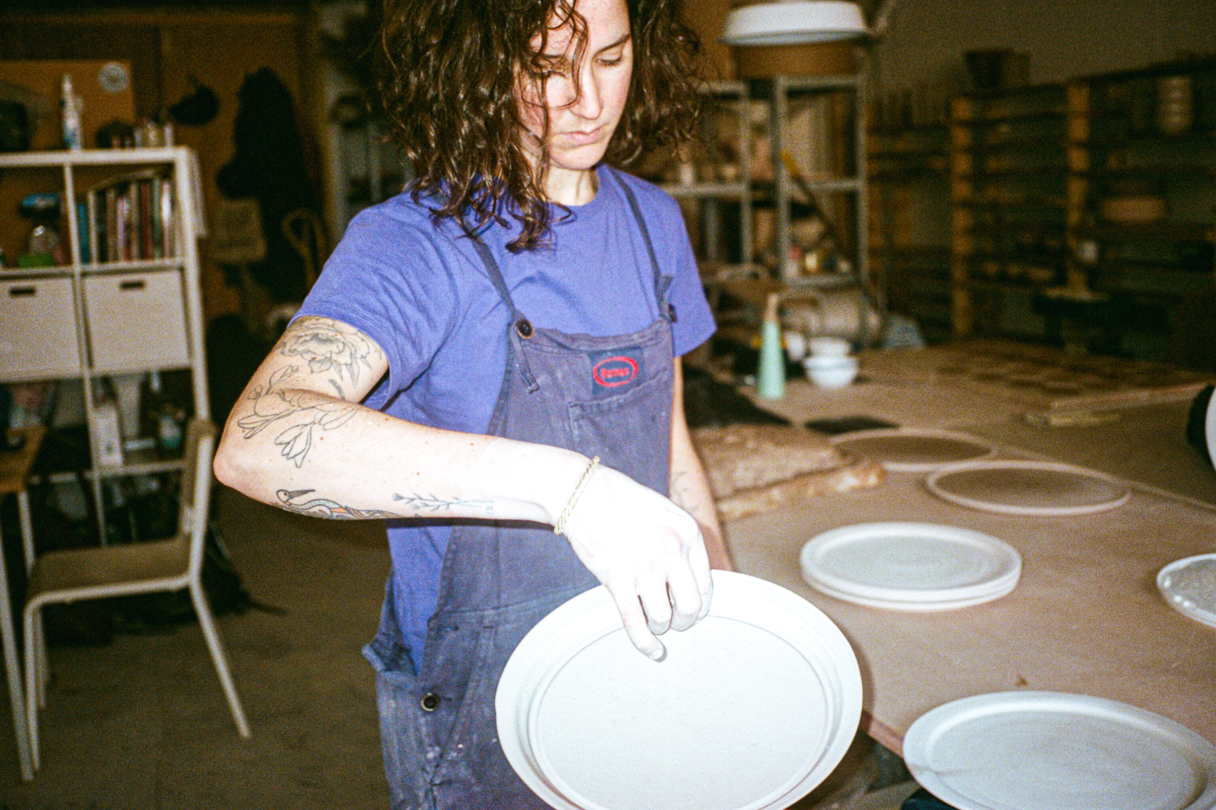 Person with tattoos and curly hair holding a white ceramic plate in a pottery studio, wearing a blue shirt and apron, surrounded by pottery items.
