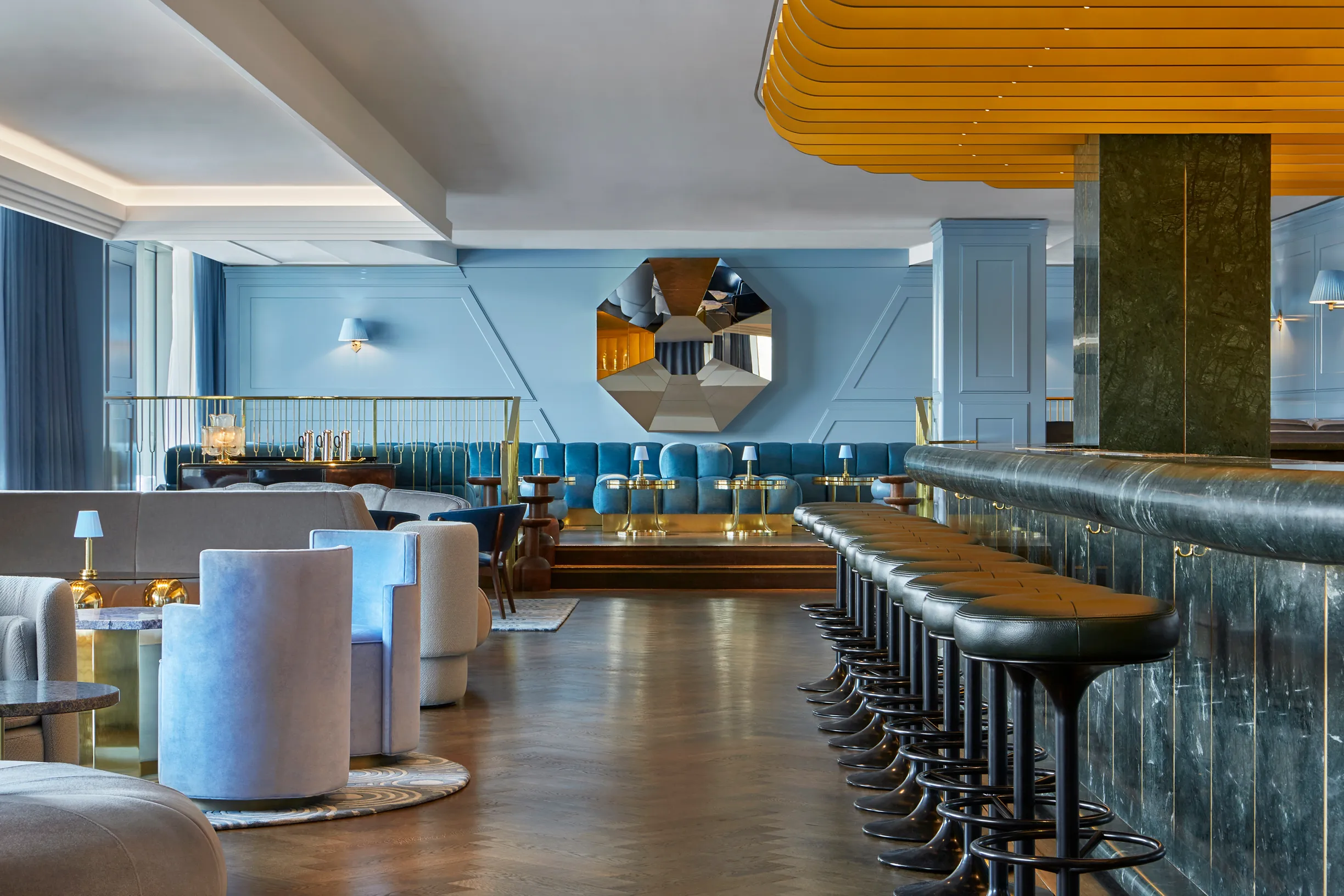 Sophisticated lounge with blue and gray seating, mirrored wall art, and a sleek bar with padded stools, featuring soft lighting and unique decor.