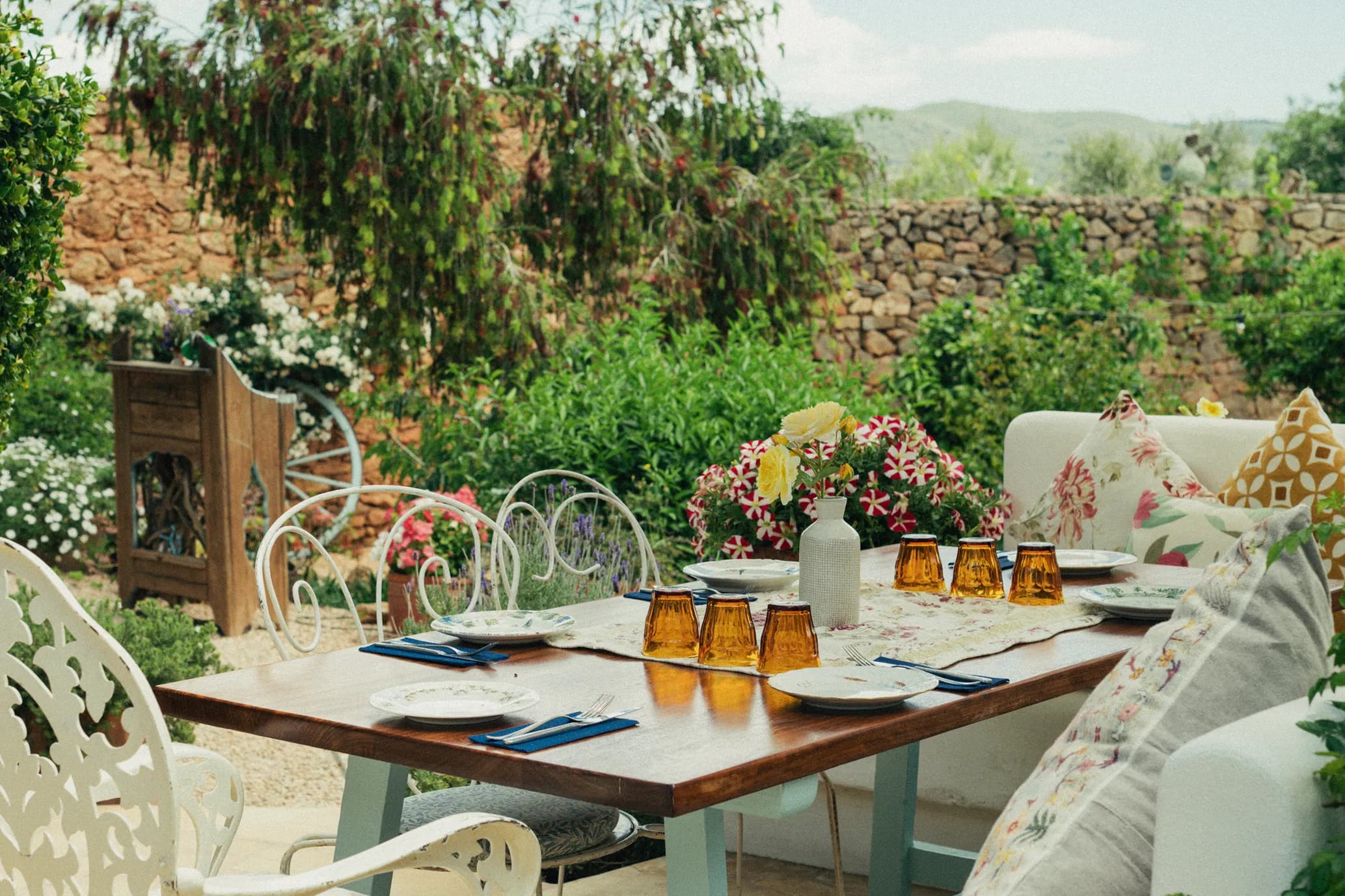 A patio dining table set with orange glasses, plates, and flowers, surrounded by decorative pillows and lush greenery in a garden setting.