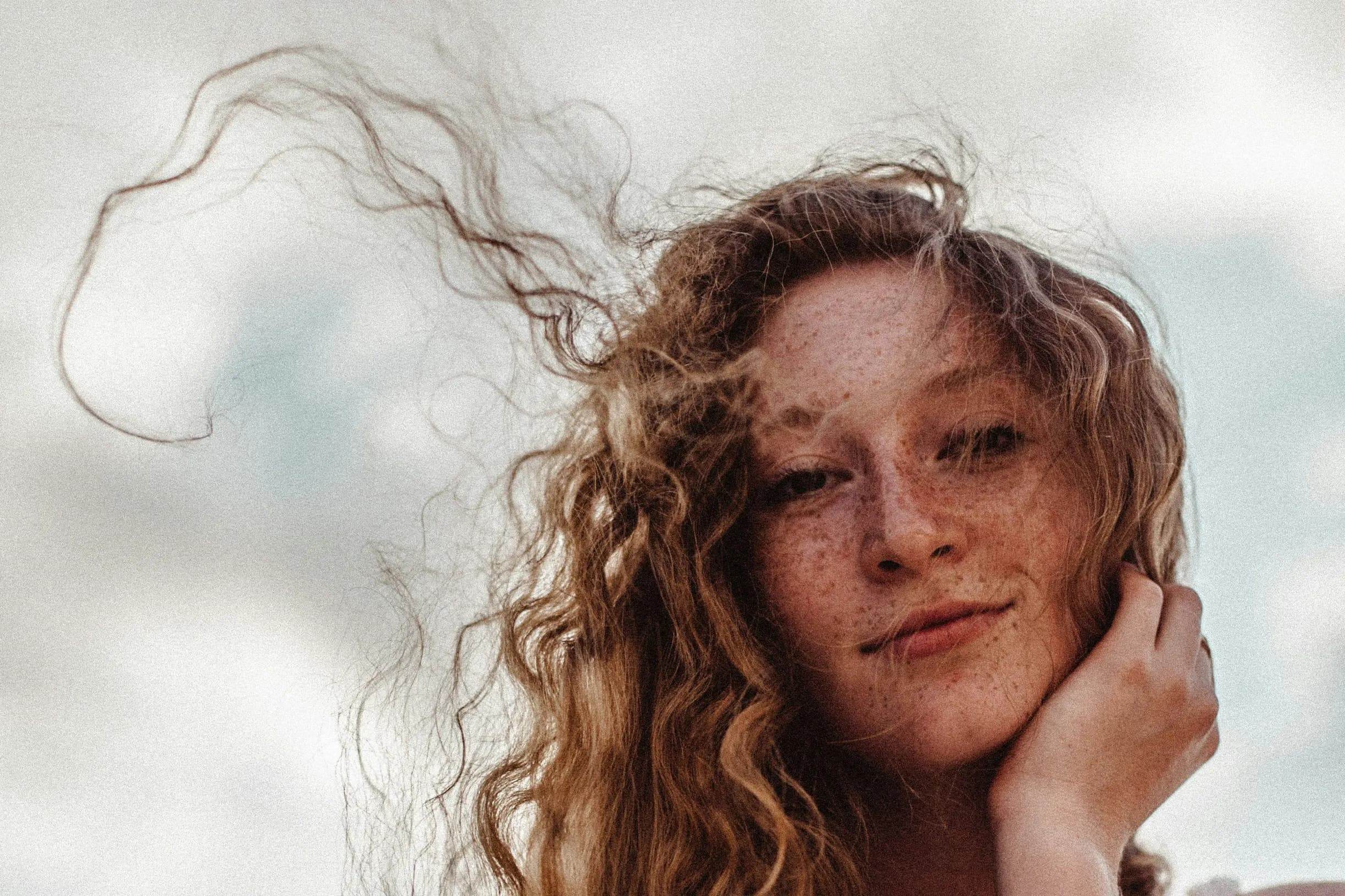 A young woman with curly, windswept hair and freckles, smiling softly against a cloudy sky background.