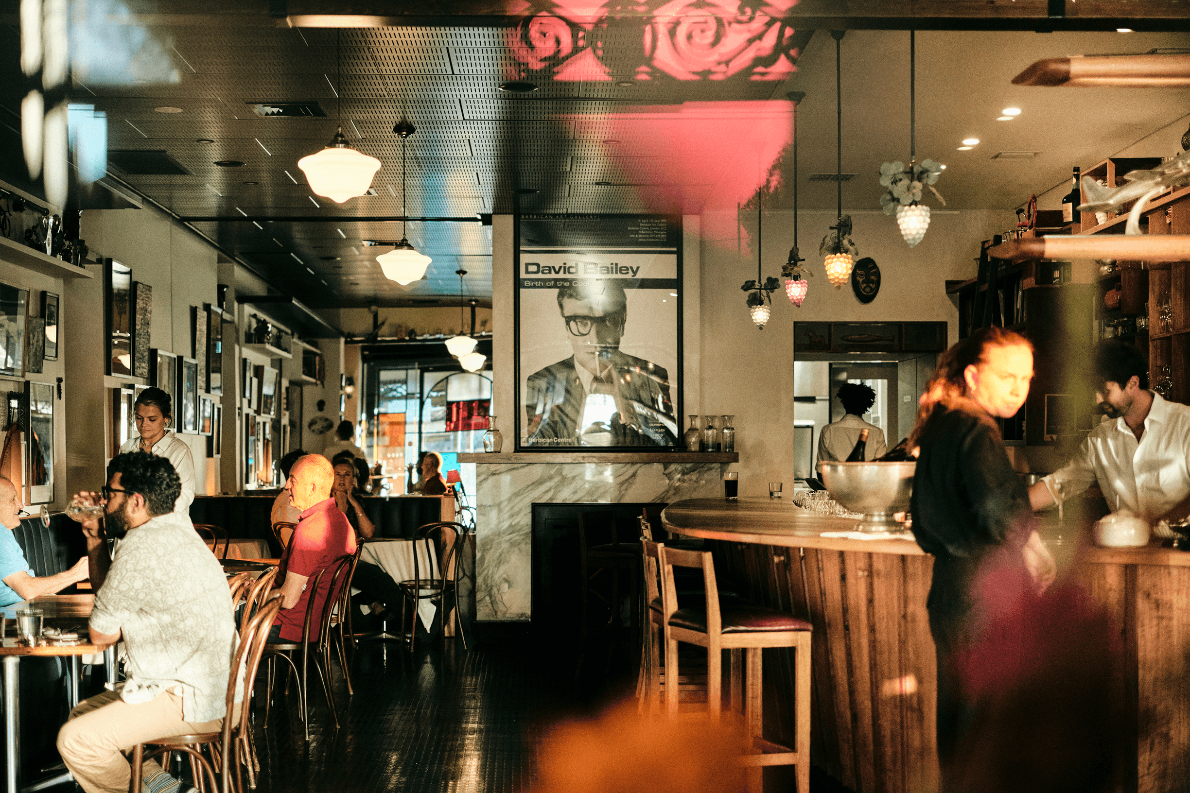 Cozy café interior with people dining and talking, warm lighting, a wooden bar, and a large portrait on the wall.