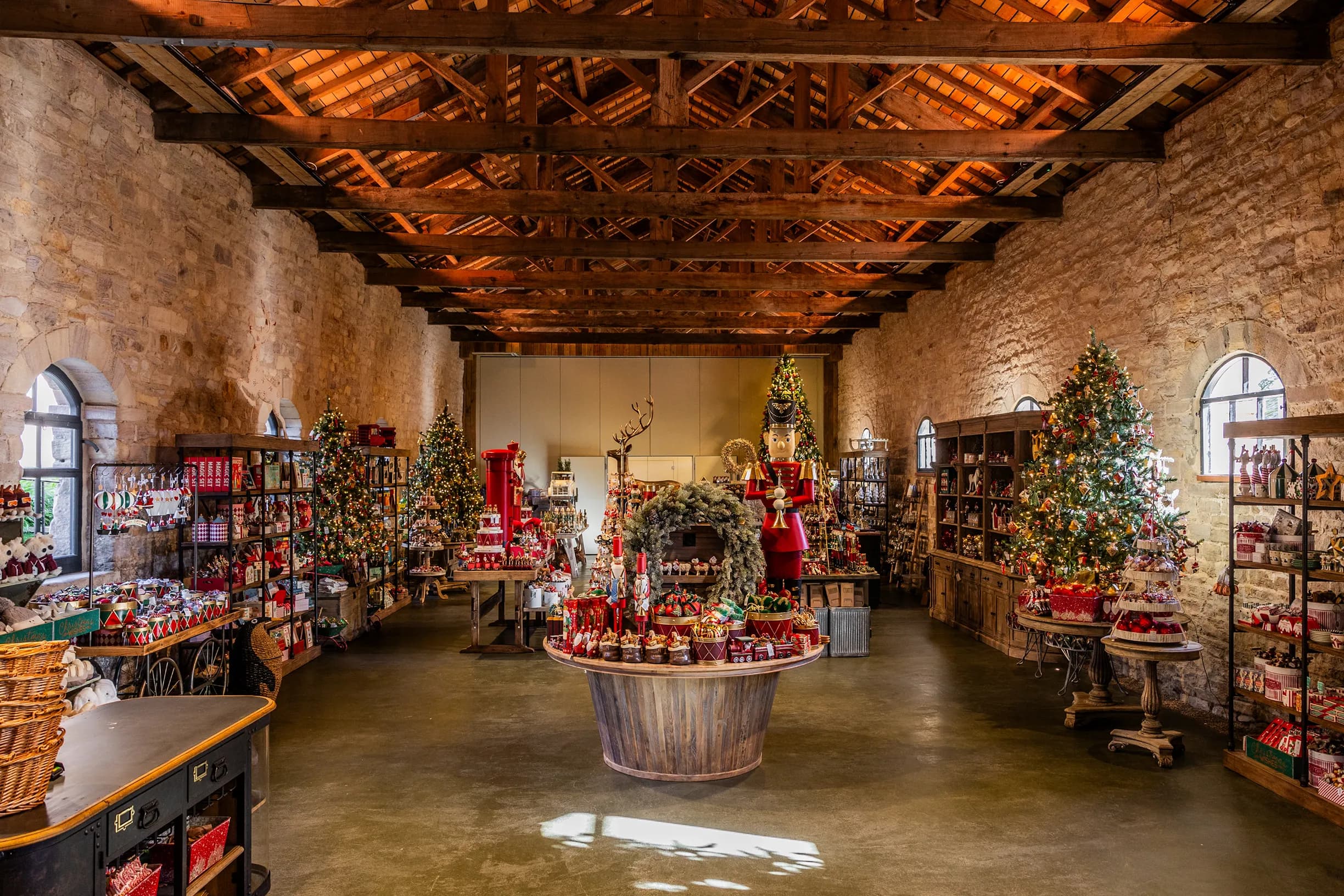 A spacious room with stone walls and wooden beams, lavishly decorated for Christmas with multiple illuminated trees, ornaments, gifts, a large nutcracker, and a red postbox.