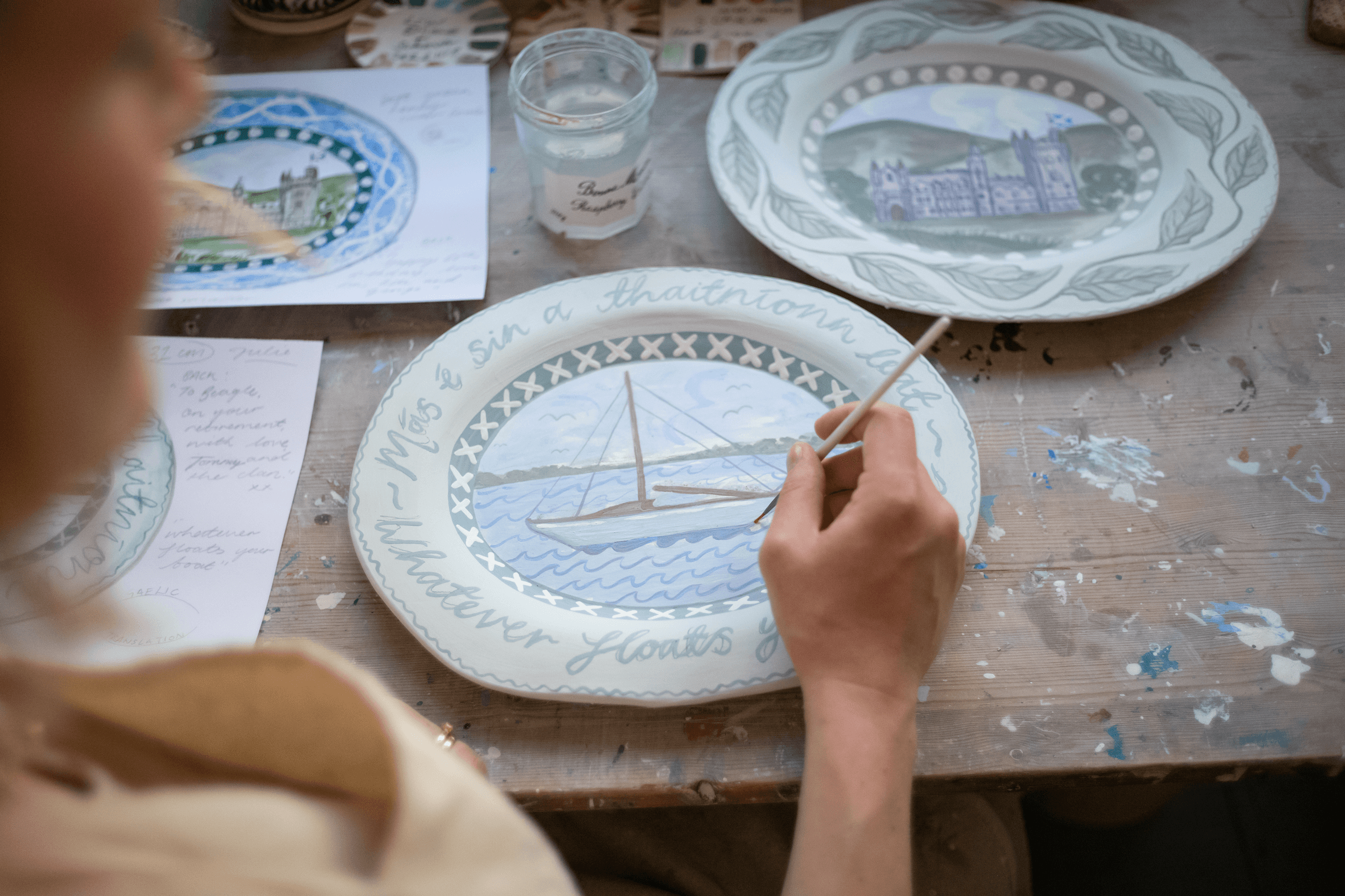 Person painting a decorative plate with a nautical scene at a wooden table, surrounded by other hand-painted plates and art supplies.