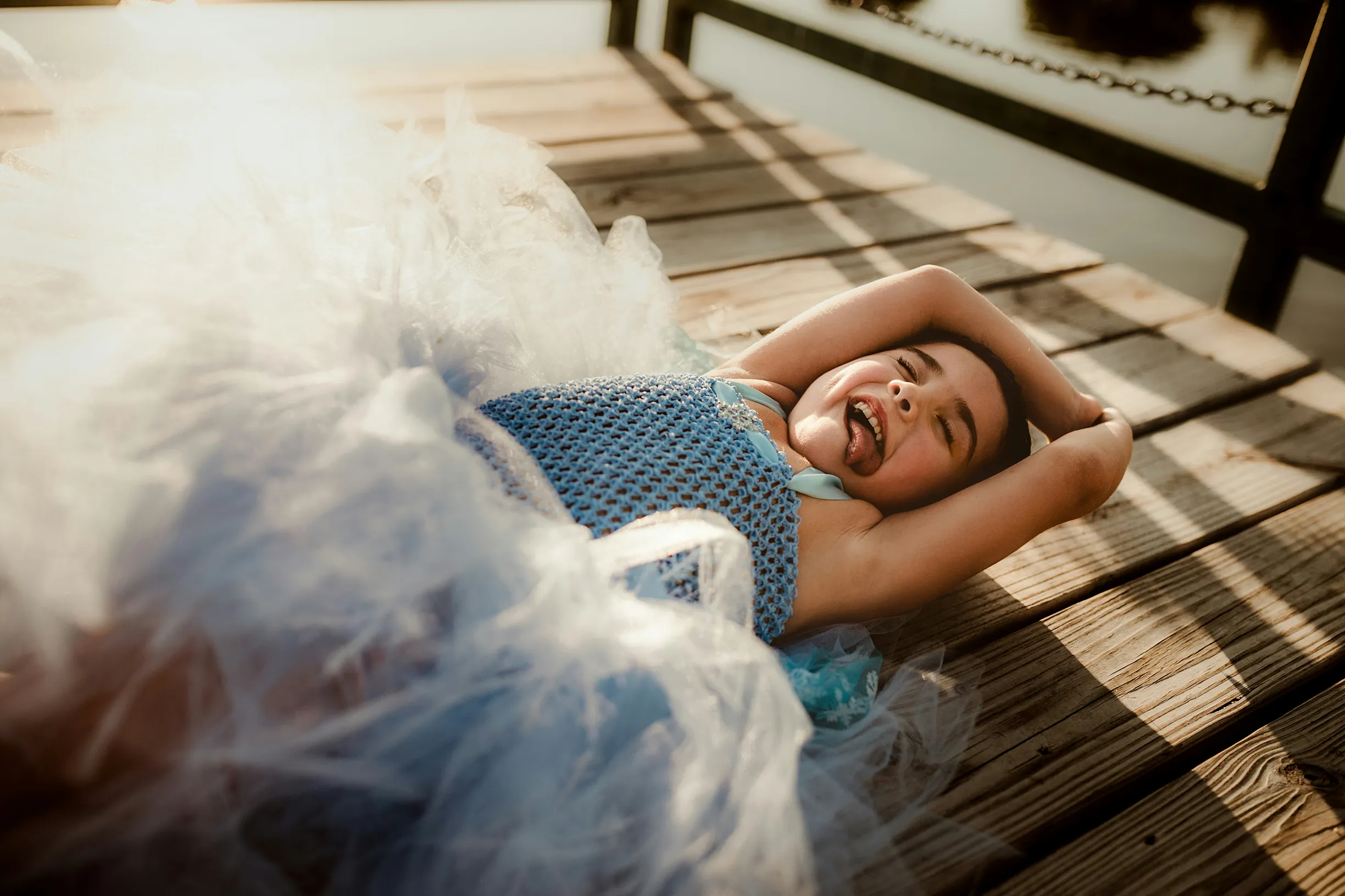Child in a blue dress with tulle, lying on a wooden pier with arms behind head, smiling in warm sunlight.
