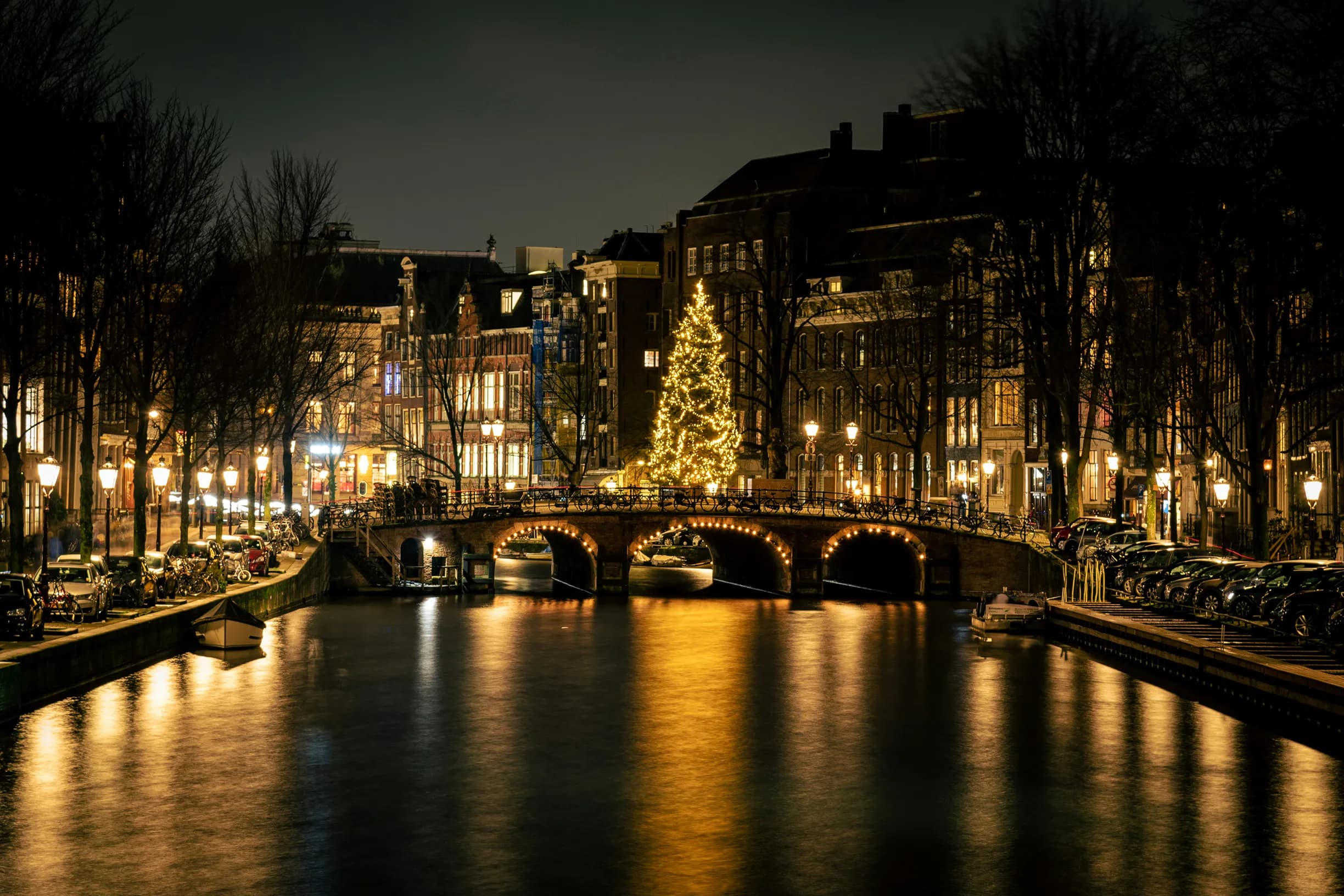 Illuminated canal in Amsterdam at night, with a lit-up Christmas tree on a bridge, reflections on the water, and buildings lining the waterway.
