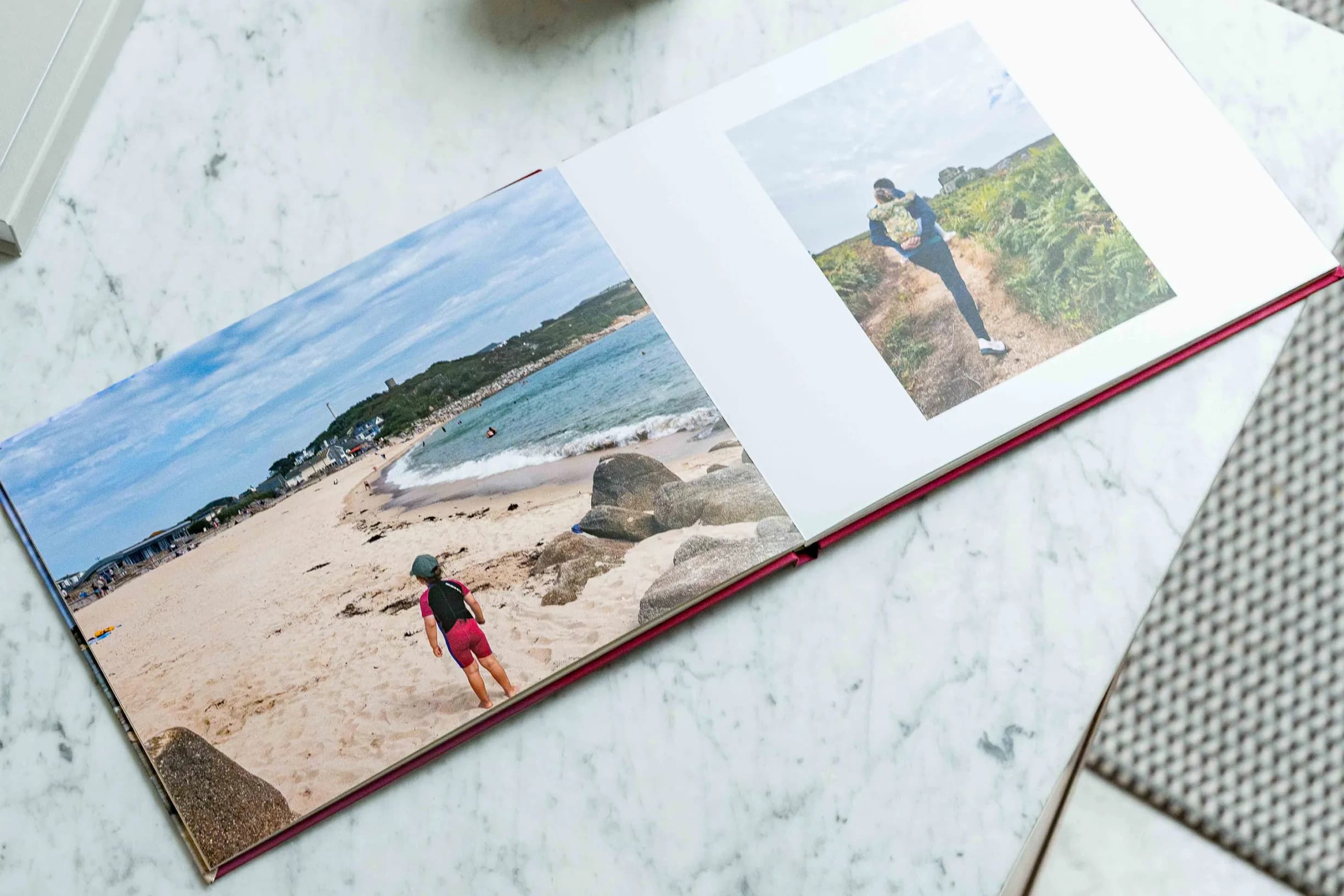 Open photo book on a marble table displaying beach and nature scenes, with a child on the beach and a person walking on a path.