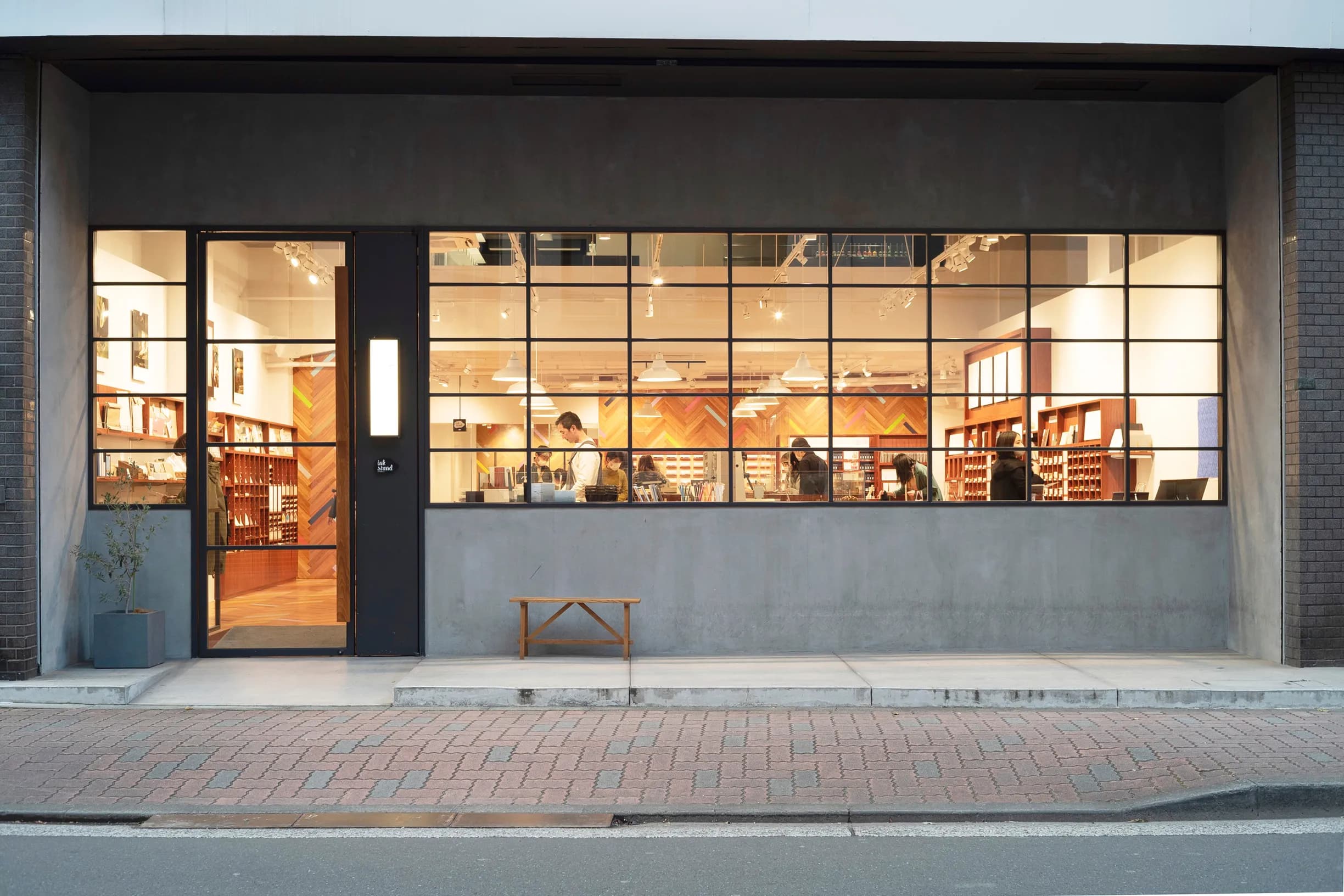 Exterior of a modern shop with large grid windows, a warmly lit interior where people are browsing, a wooden bench, and a brick sidewalk.