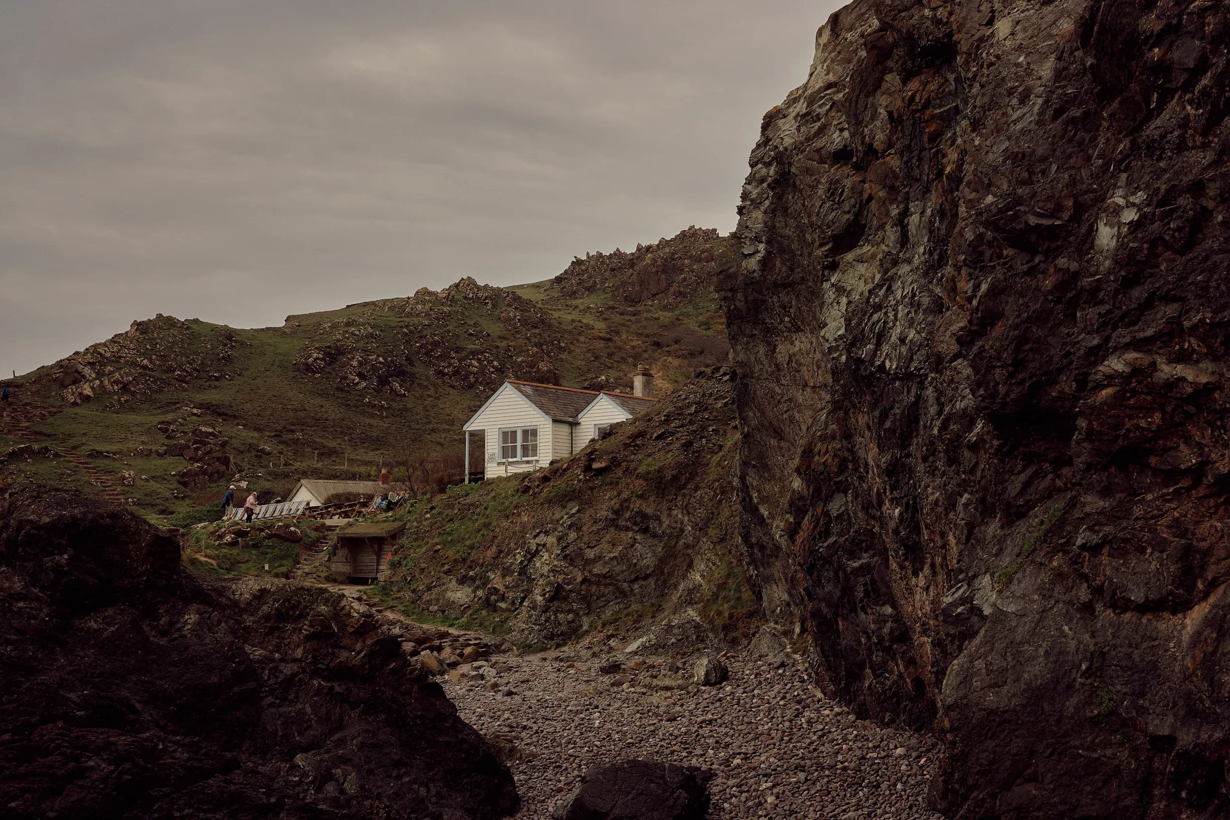 A white house beside rugged cliffs and grassy hills, with a pebble beach in the foreground under an overcast sky.