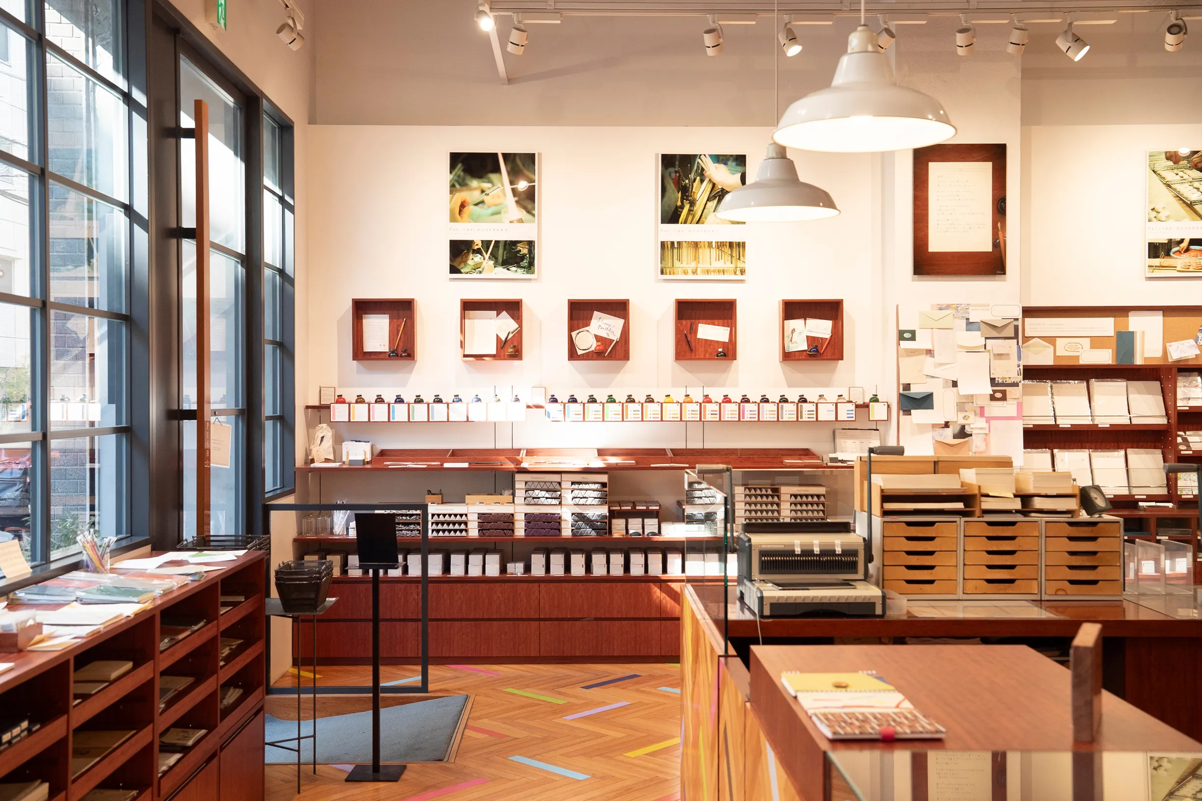 Interior of a modern stationery store with large windows, wooden displays of ink, paper, and pens, artwork, and a herringbone floor with colorful stripes.