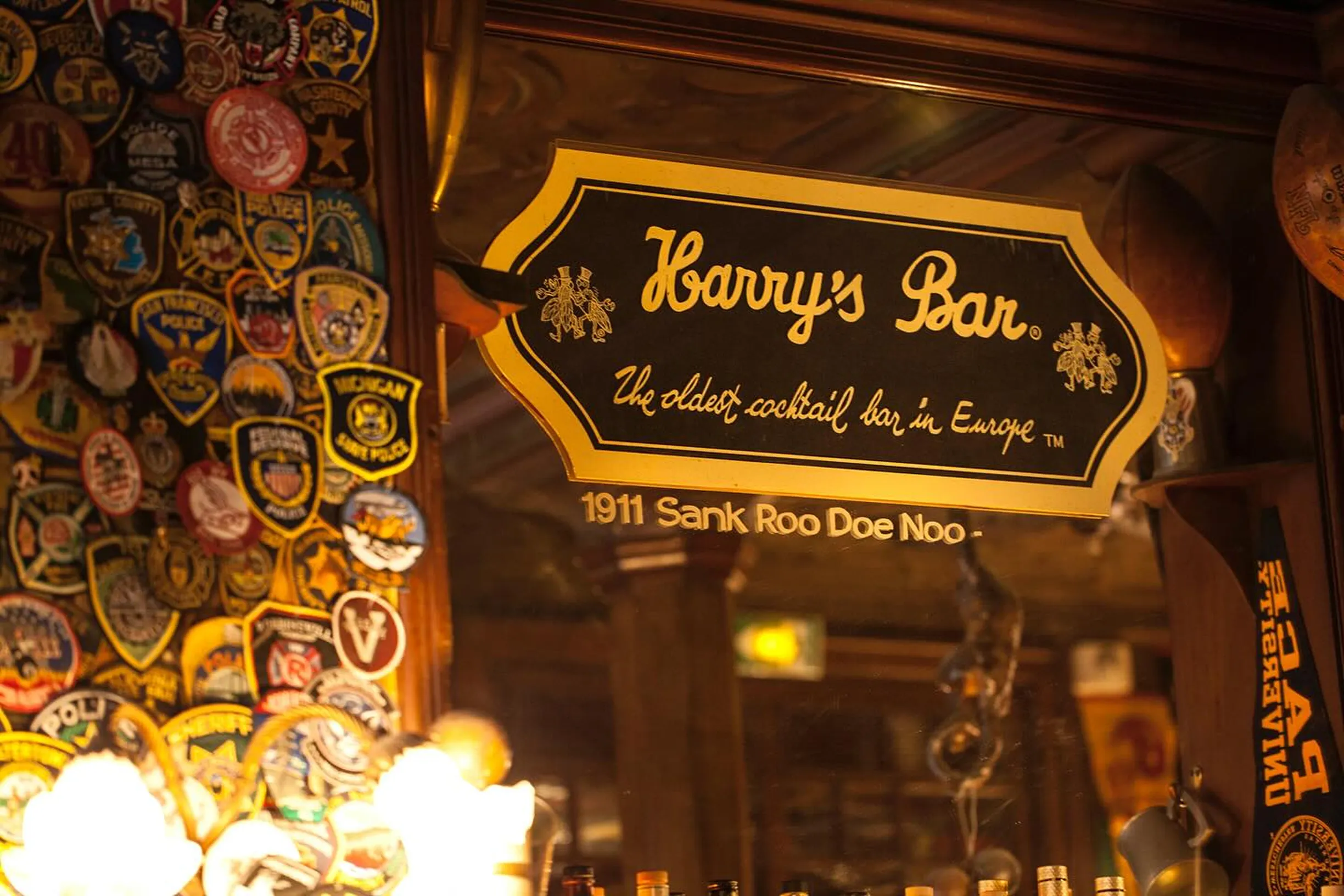 Sign reading "Harry's Bar, the oldest cocktail bar in Europe," surrounded by various patches and memorabilia.