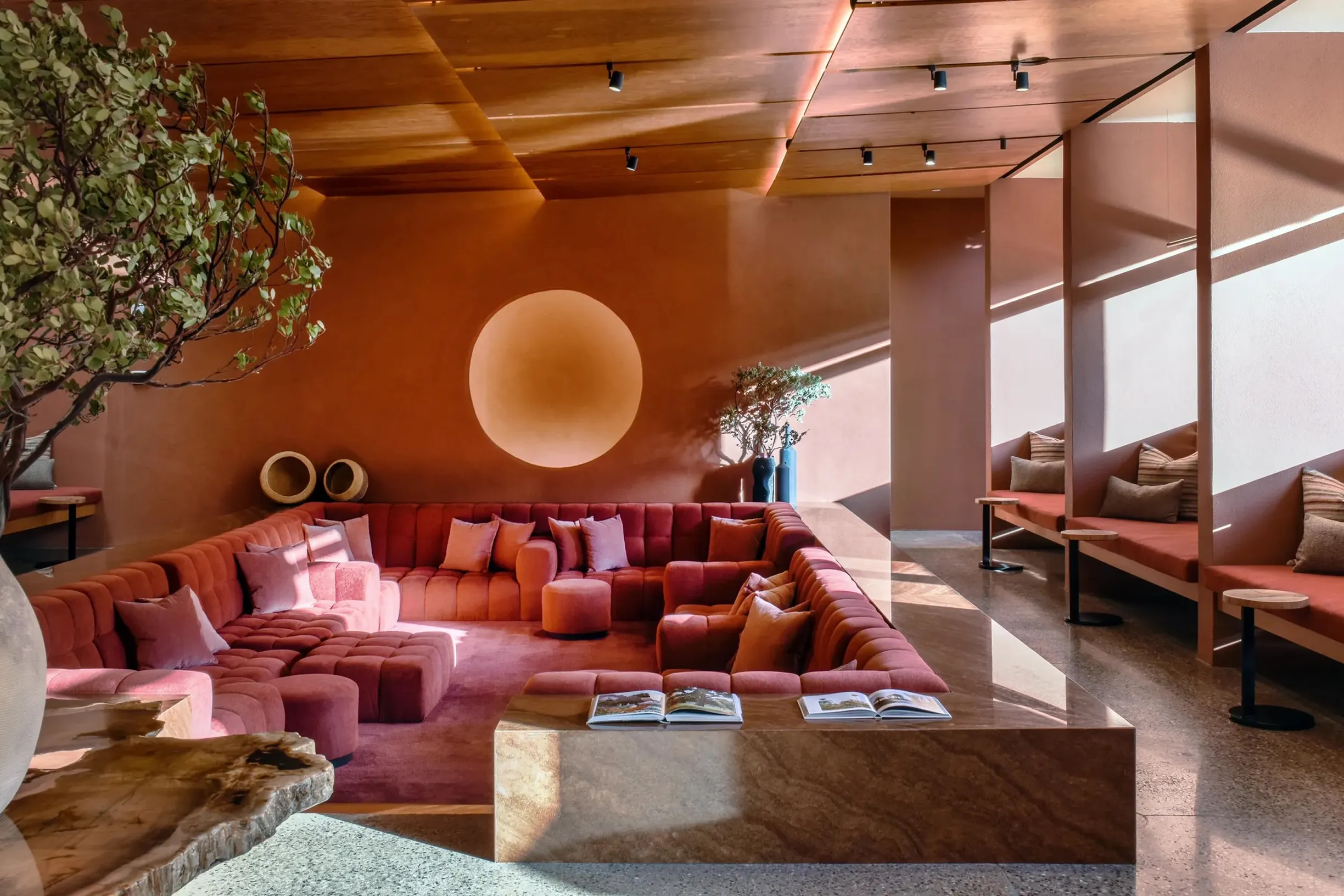 Stylish modern lobby in warm terracotta and rose hues, featuring a large U-shaped sofa, circular wall light, and sunlit individual lounge booths.