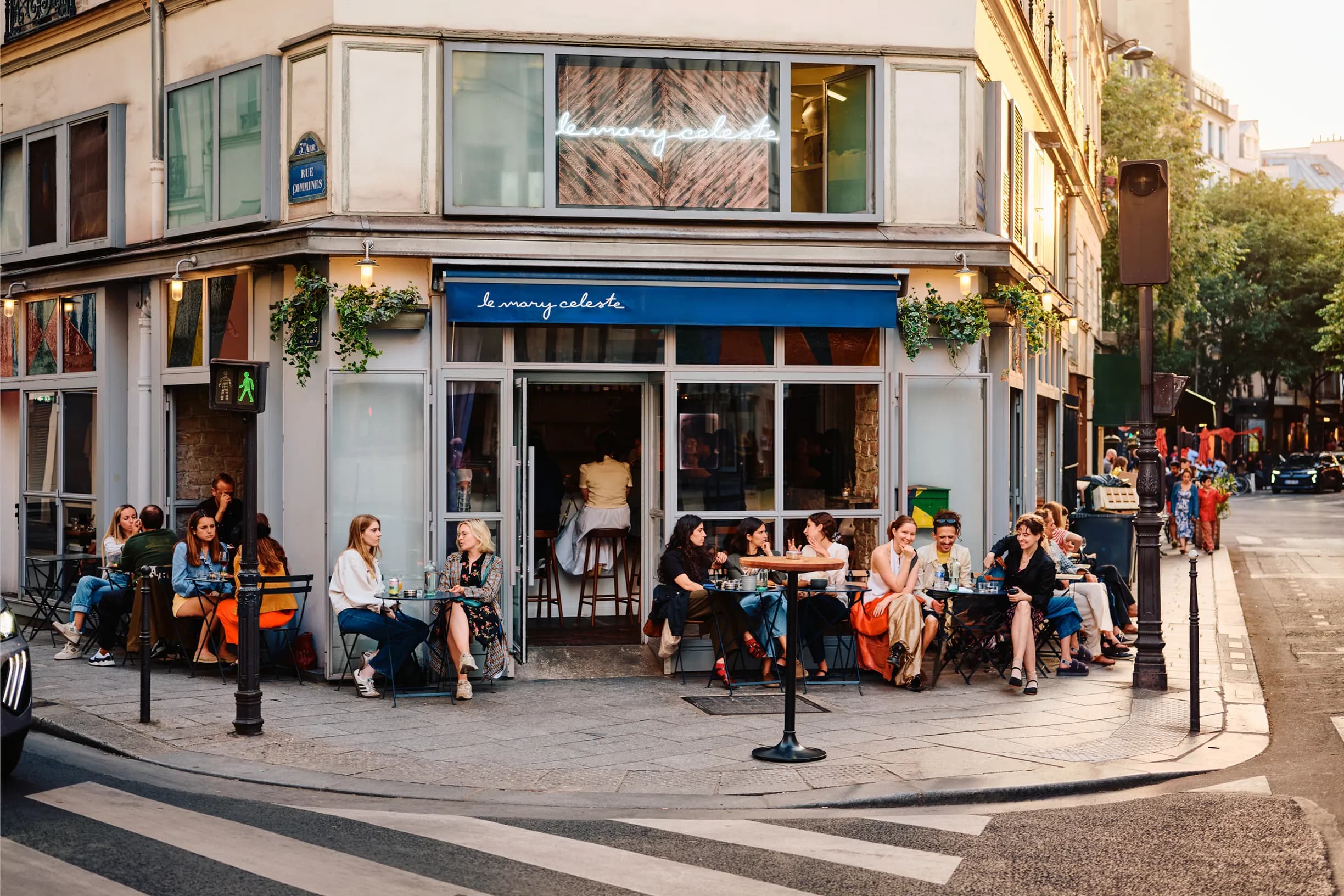 People sitting at outdoor tables of a corner café with blue awning, chatting and enjoying drinks on a sunny day.