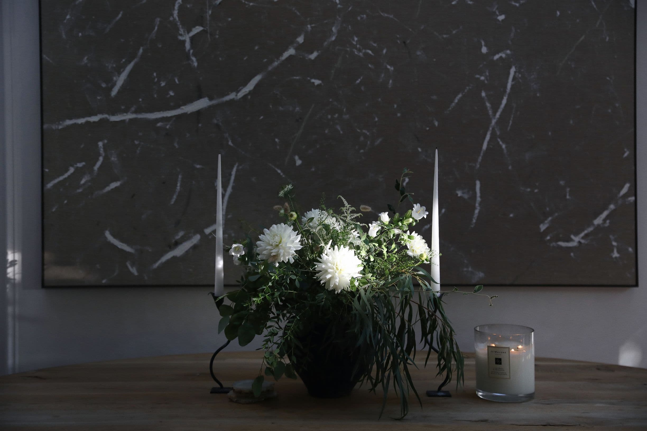 A floral arrangement with white flowers and greenery sits on a wooden table, flanked by two tall candles. A lit candle is nearby.