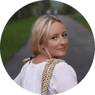 Woman in a white shirt and with a woven bag over her shoulder walking on a path, looking back, surrounded by greenery.