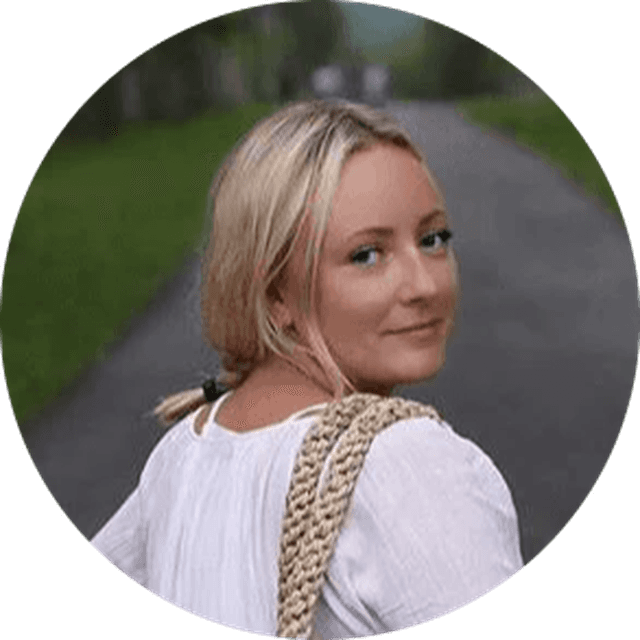 Woman in a white shirt and with a woven bag over her shoulder walking on a path, looking back, surrounded by greenery.