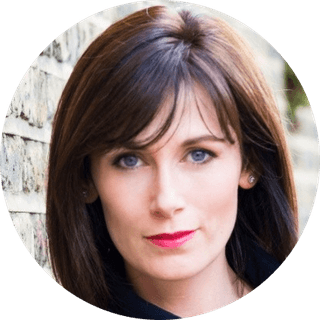 Close headshot of a woman with shoulder-length brown hair, blue eyes, fair skin and pink lipstick, posed against a brick wall.
