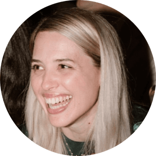 A person with long blonde hair smiling brightly, wearing a dark top, against a blurred background.